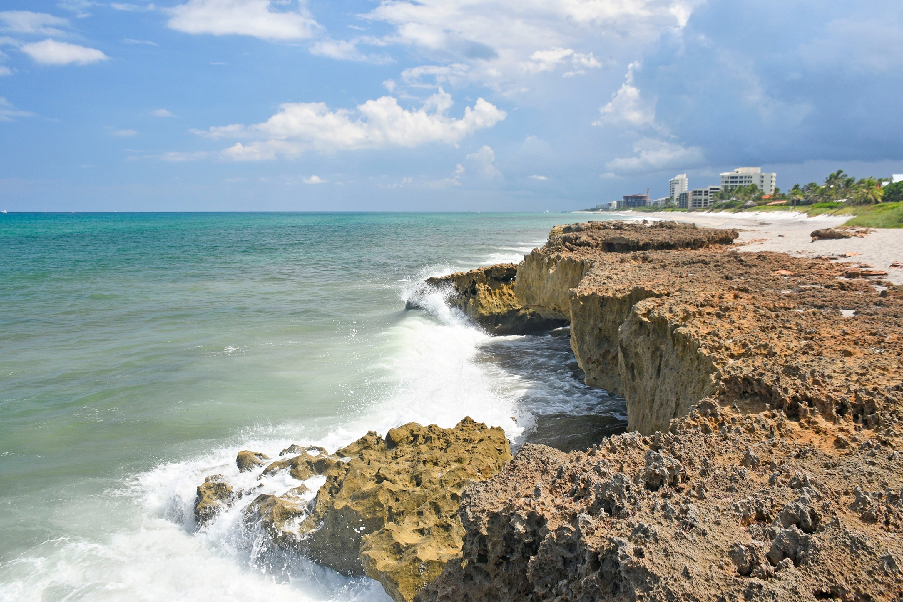Waves crash against light brown cliffs overlooking a clear blue ocean with a few buildings in the distance.