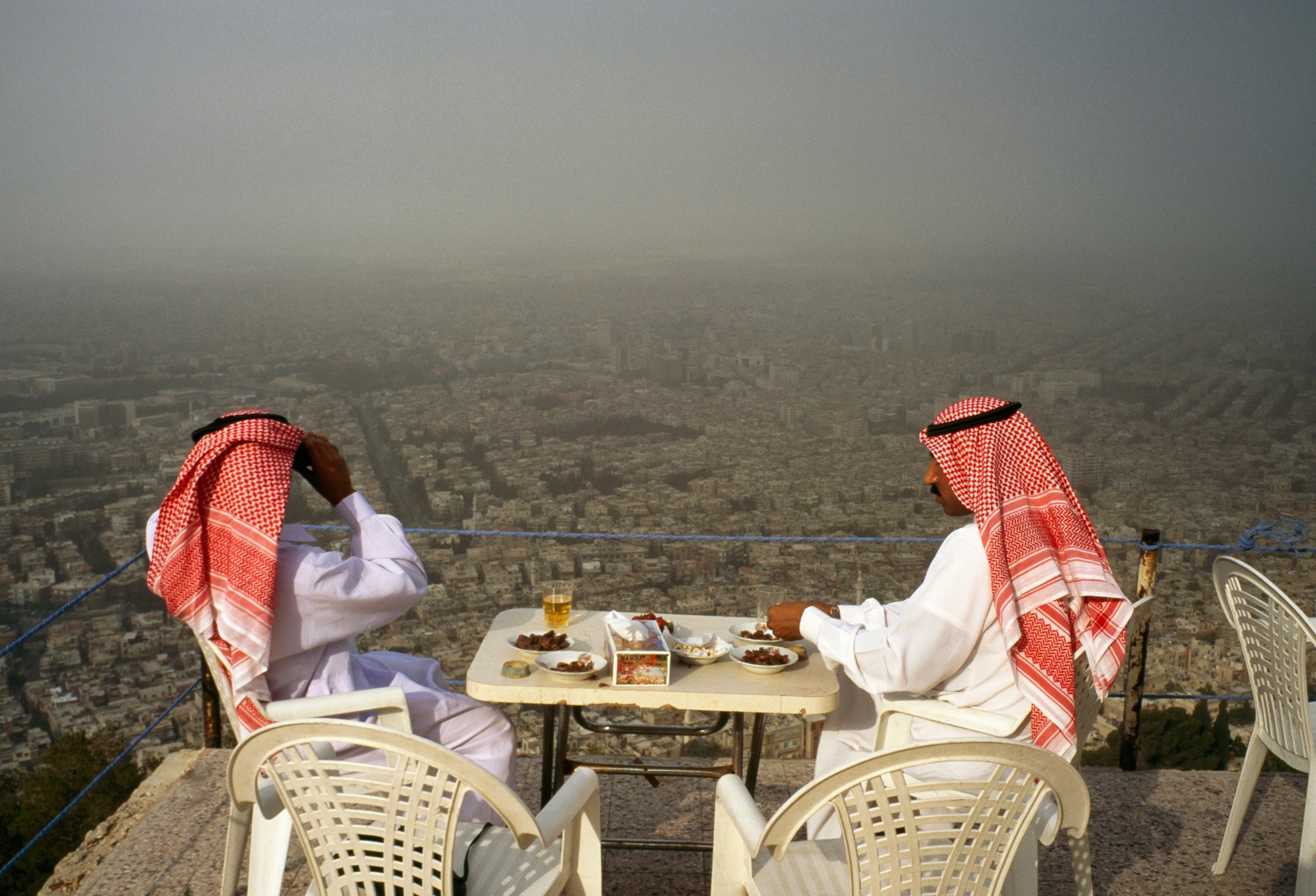 Two men in white robes and red-and-white-check headscarves sit in a cafe overlooking Damascus, 1996