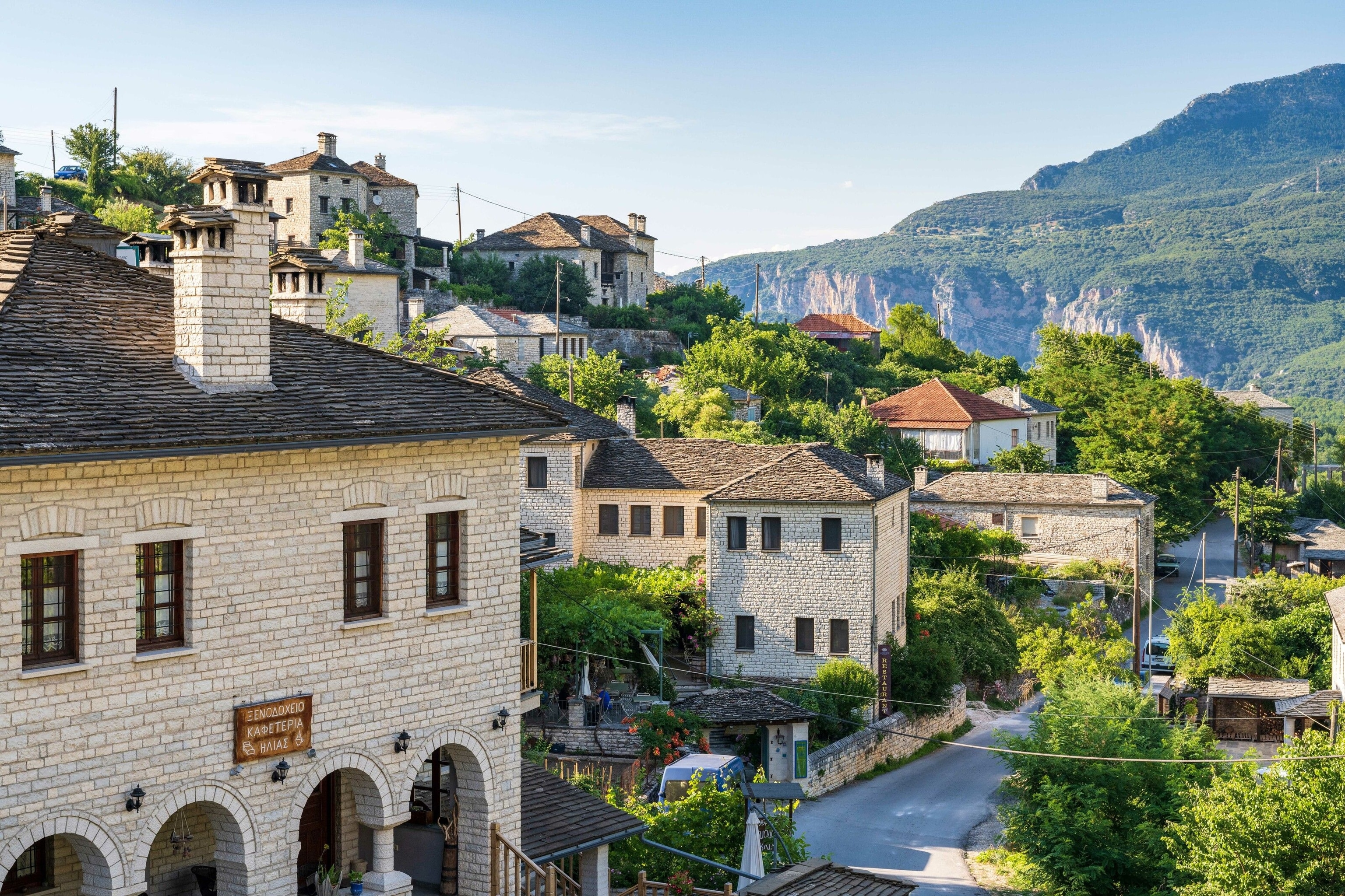 The stone buildings of Aristi. Green land and rocky cliffs are in the background, caught in the sun.