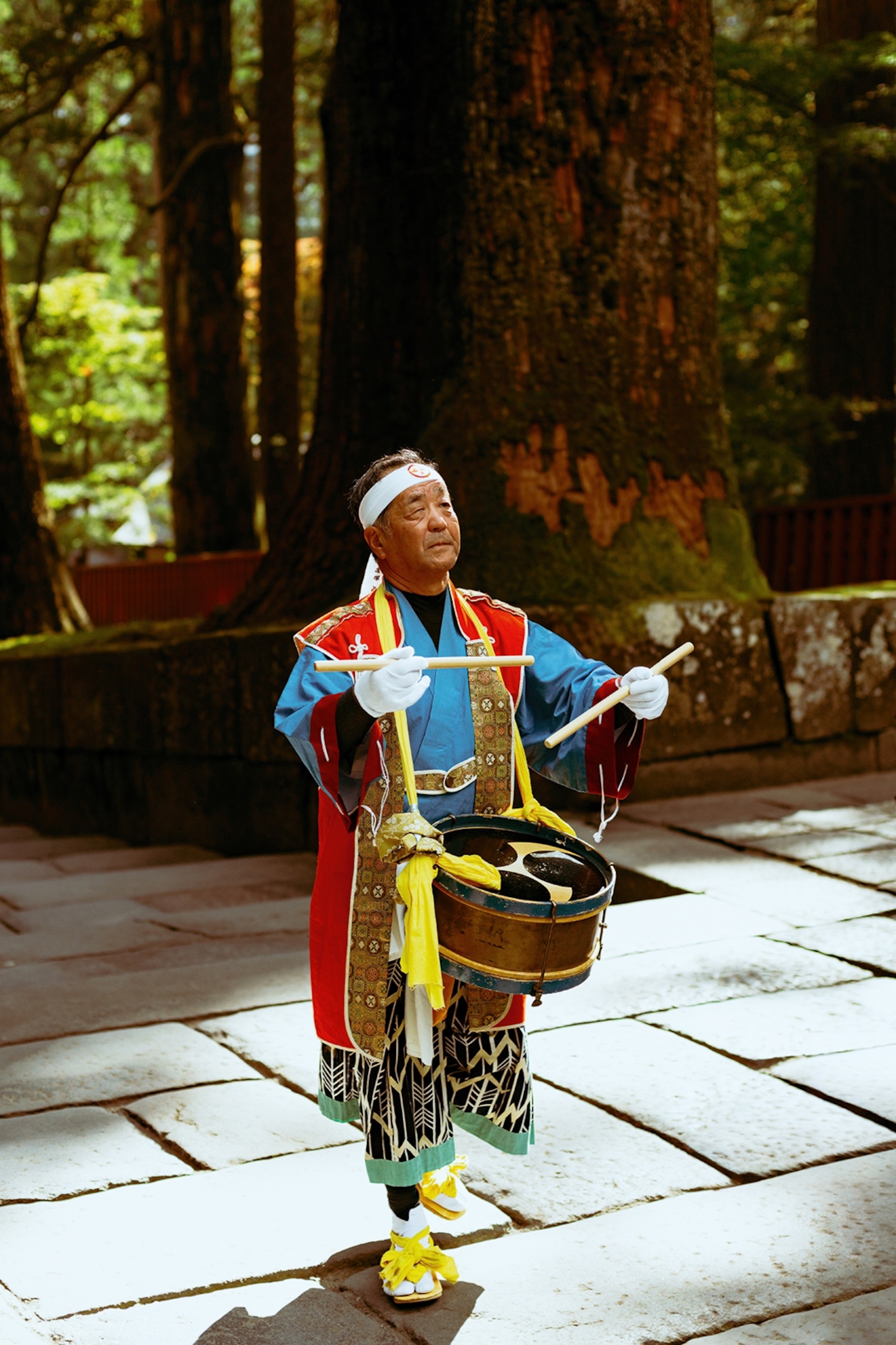 An elderly Japanese man dressed in a traditional, colourful kimono playing the drums.