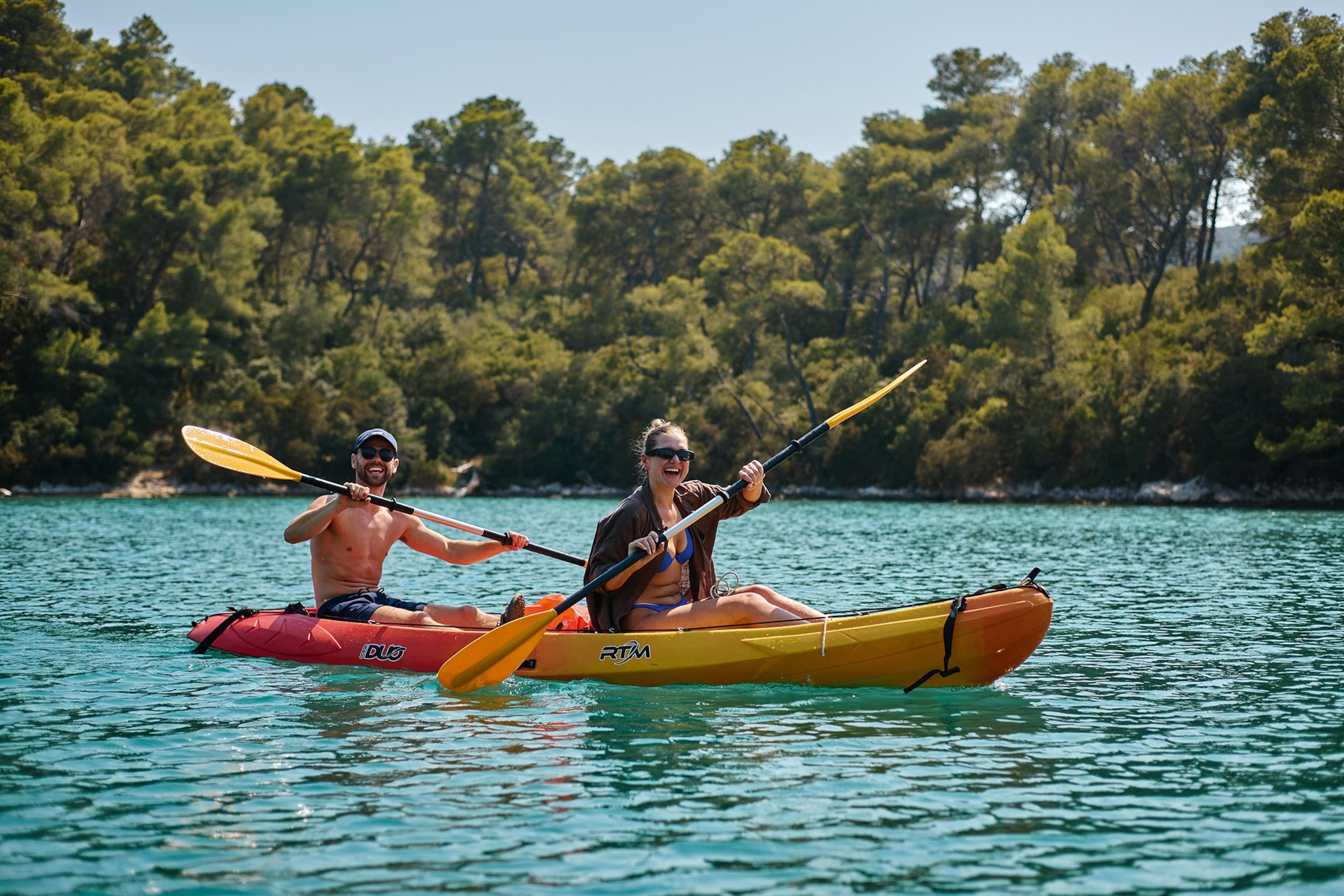 A smiling man and woman paddling down a wide river or lake in shorts and bikini in kayaks.