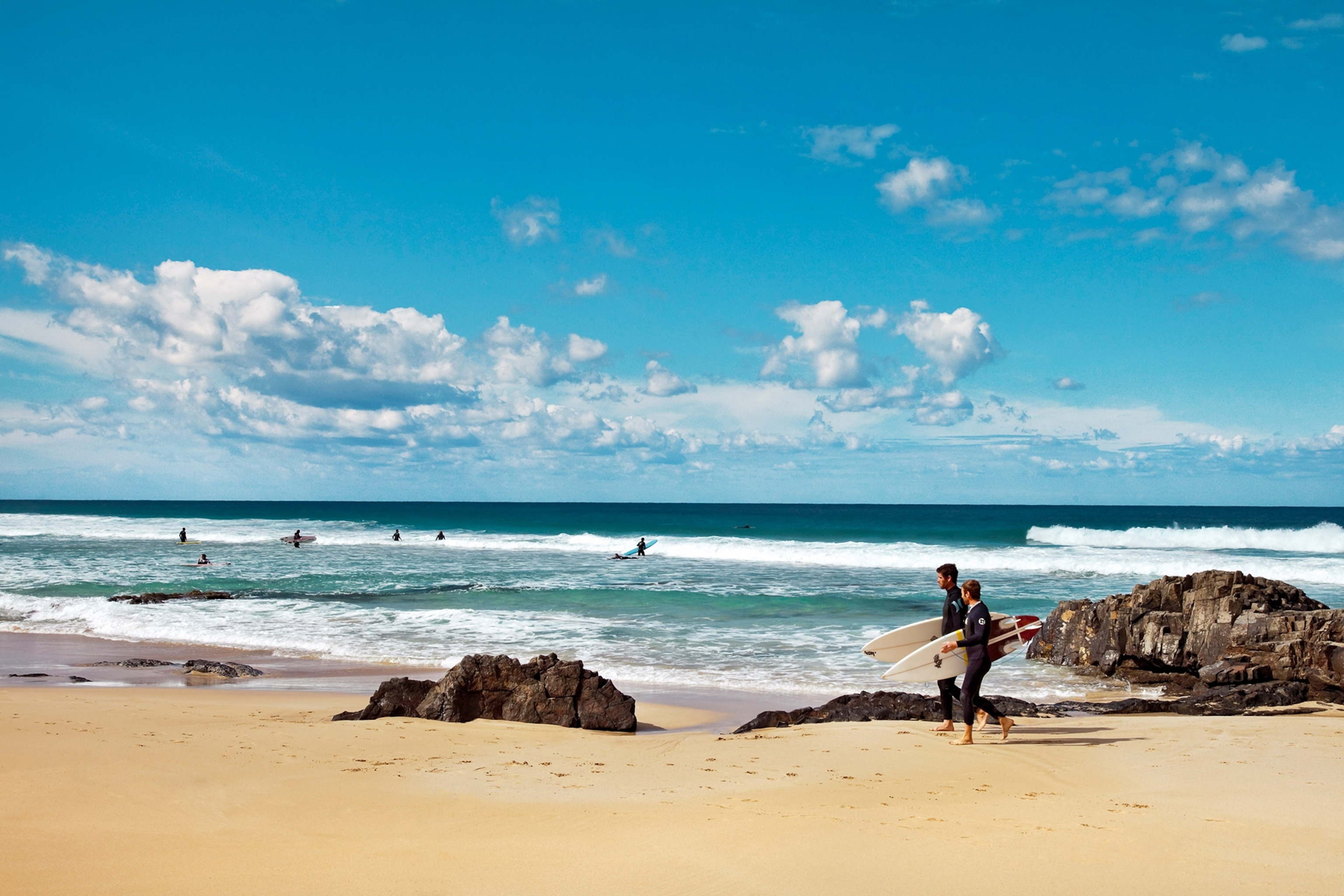 A picture-perfect beach with a long walk into the ocean as two surfers with boards pass by.
