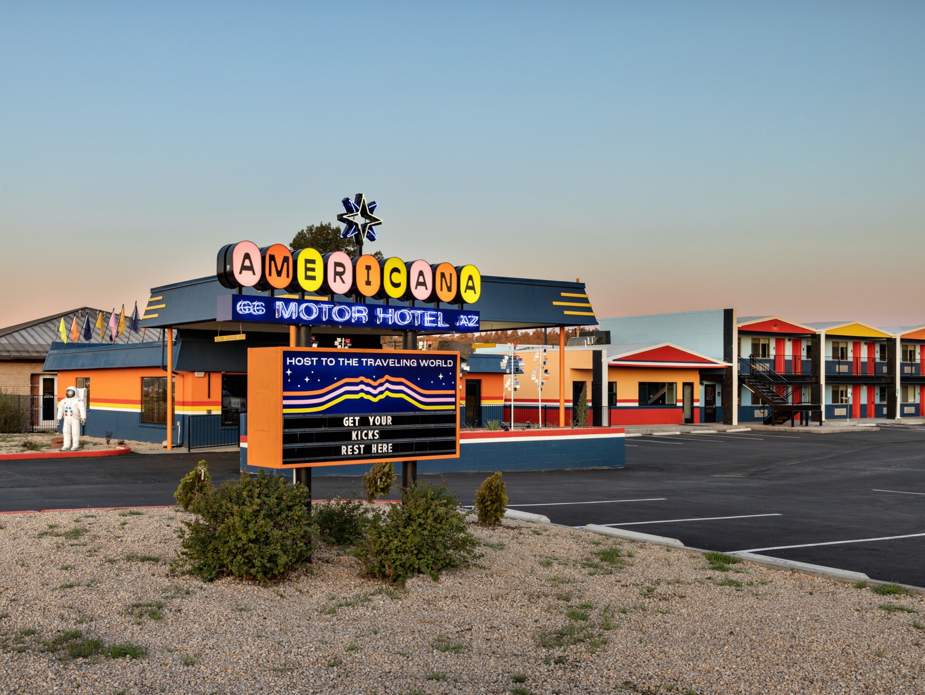 The exterior of a motor hotel and parking lot in the desert at sunset with a retro sign welcoming guests.