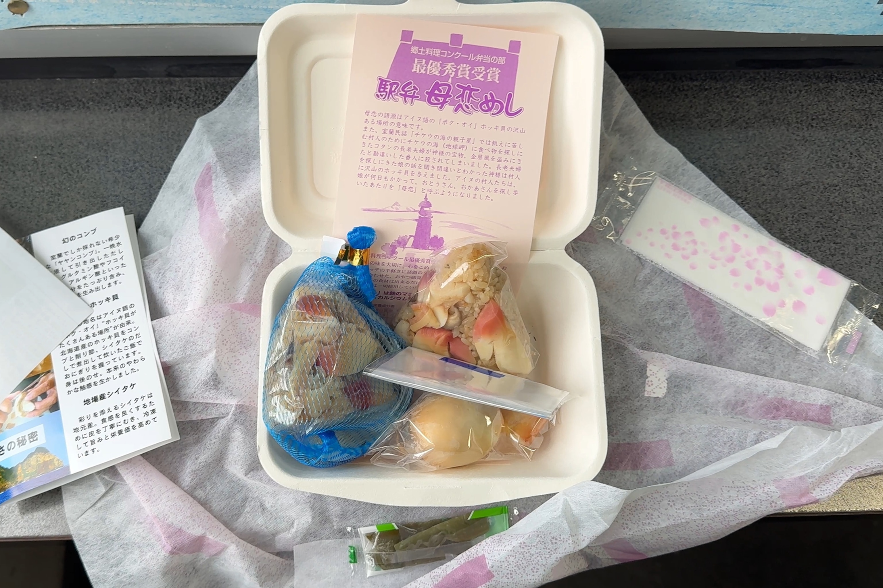 A train lunch box, unwrapped on a train table featuring clam, rice balls and tiny fish.