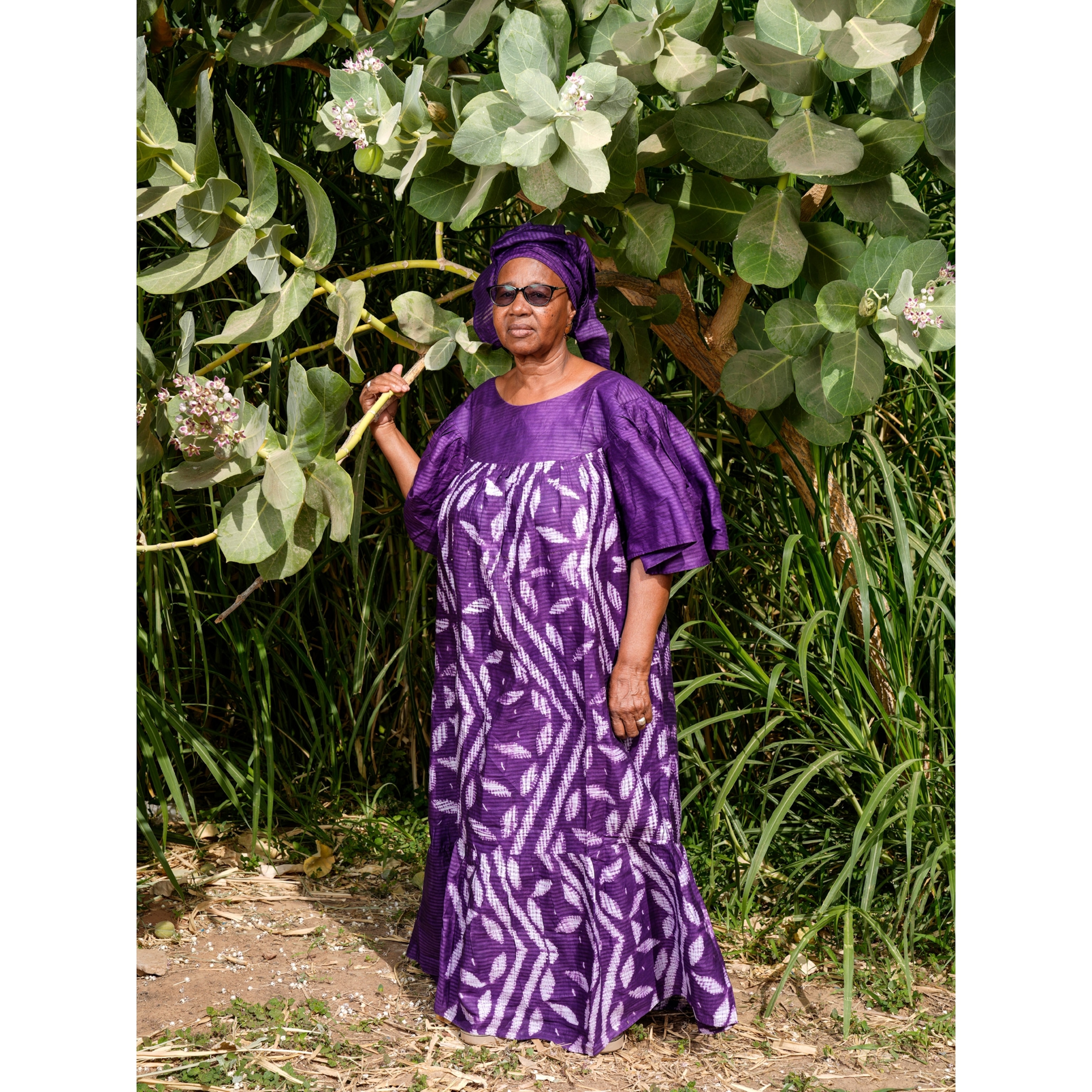 A woman stands outdoors for a portrait while wearing a purple gown, purple hair wrap, and sunglasses.