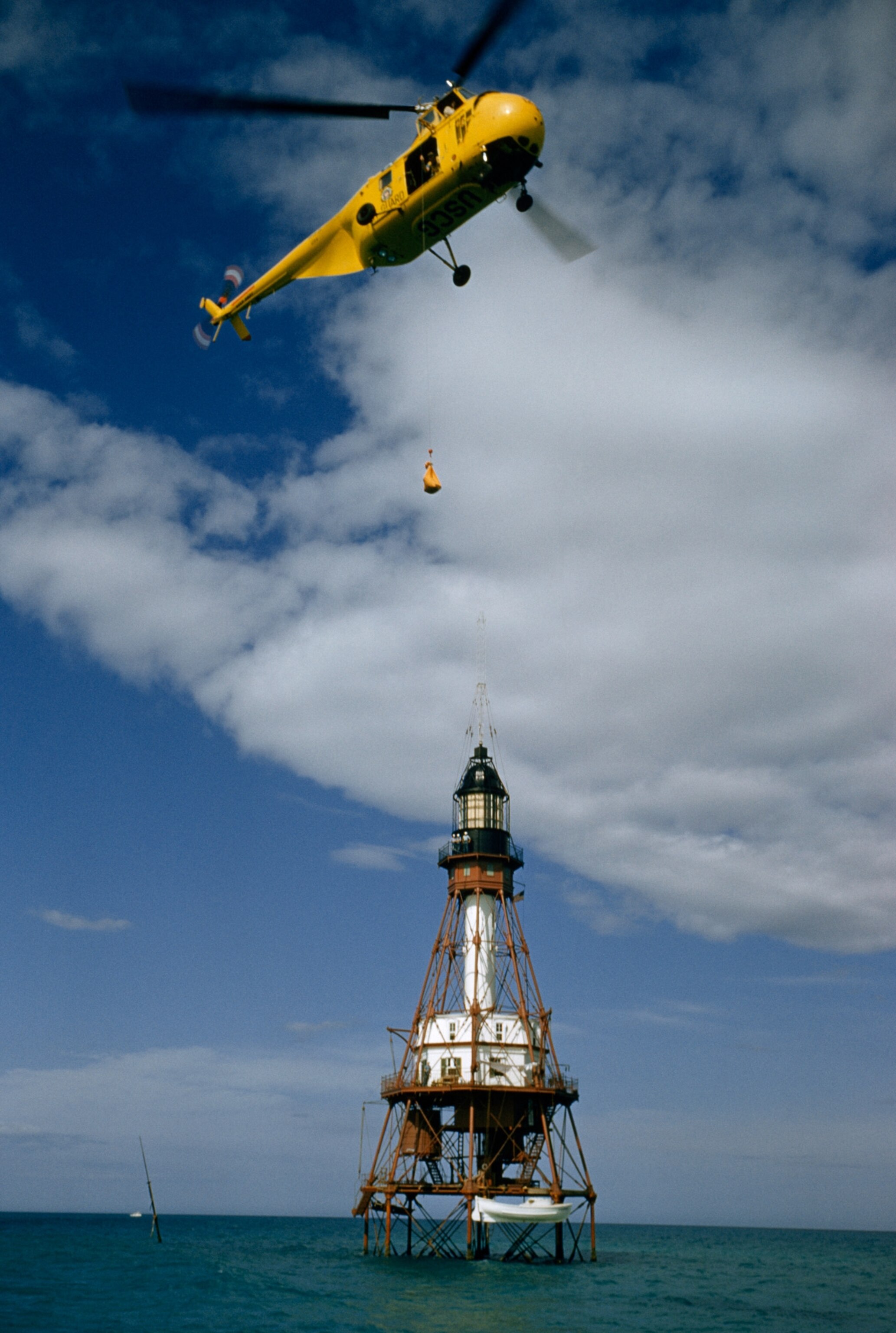 a Coast Guard helicopter dropping mail over a lighthouse at sea.