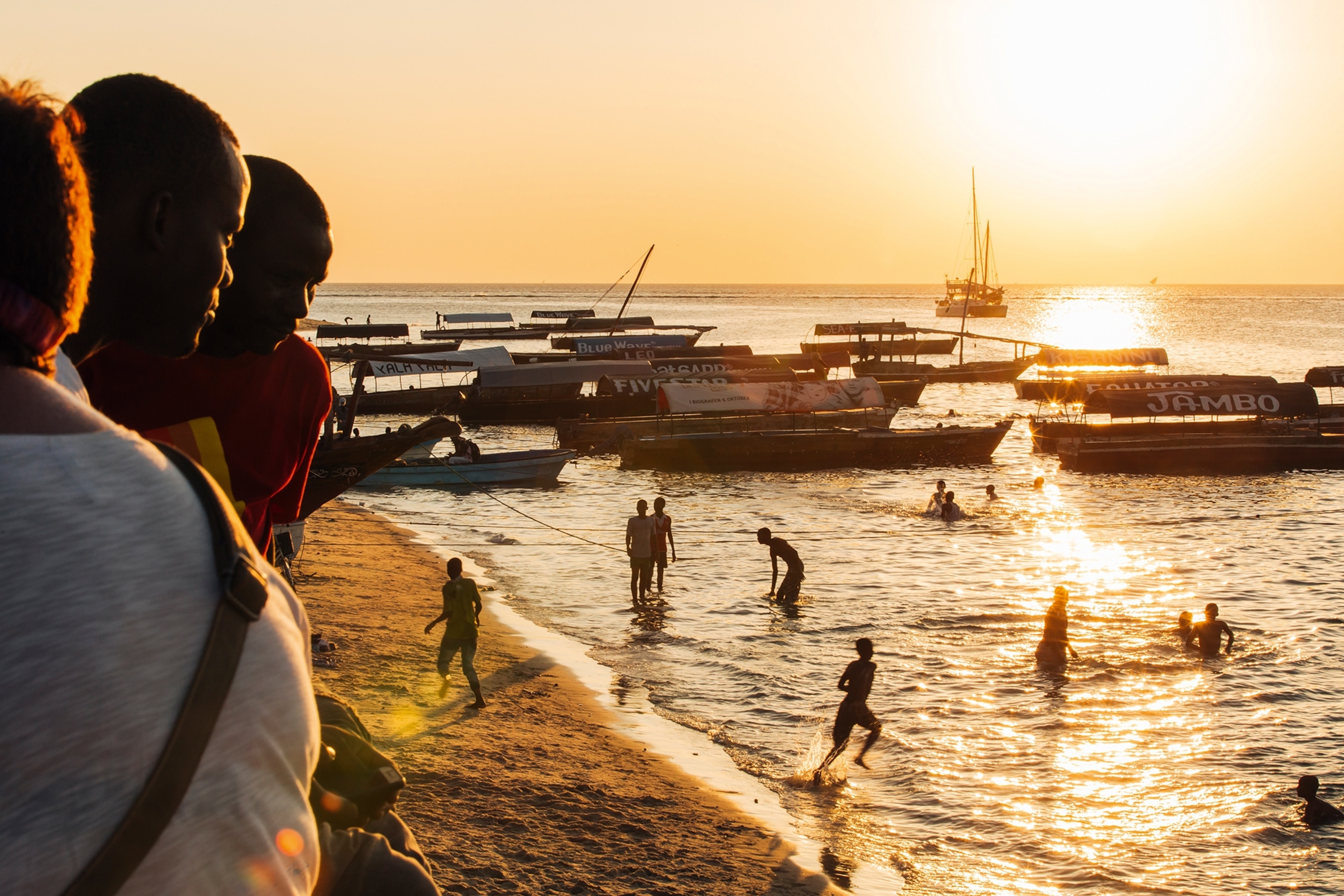 An evening sun shot of locals playing at a beach in Stone Town, Zanzibar, with boats anchored in the background.