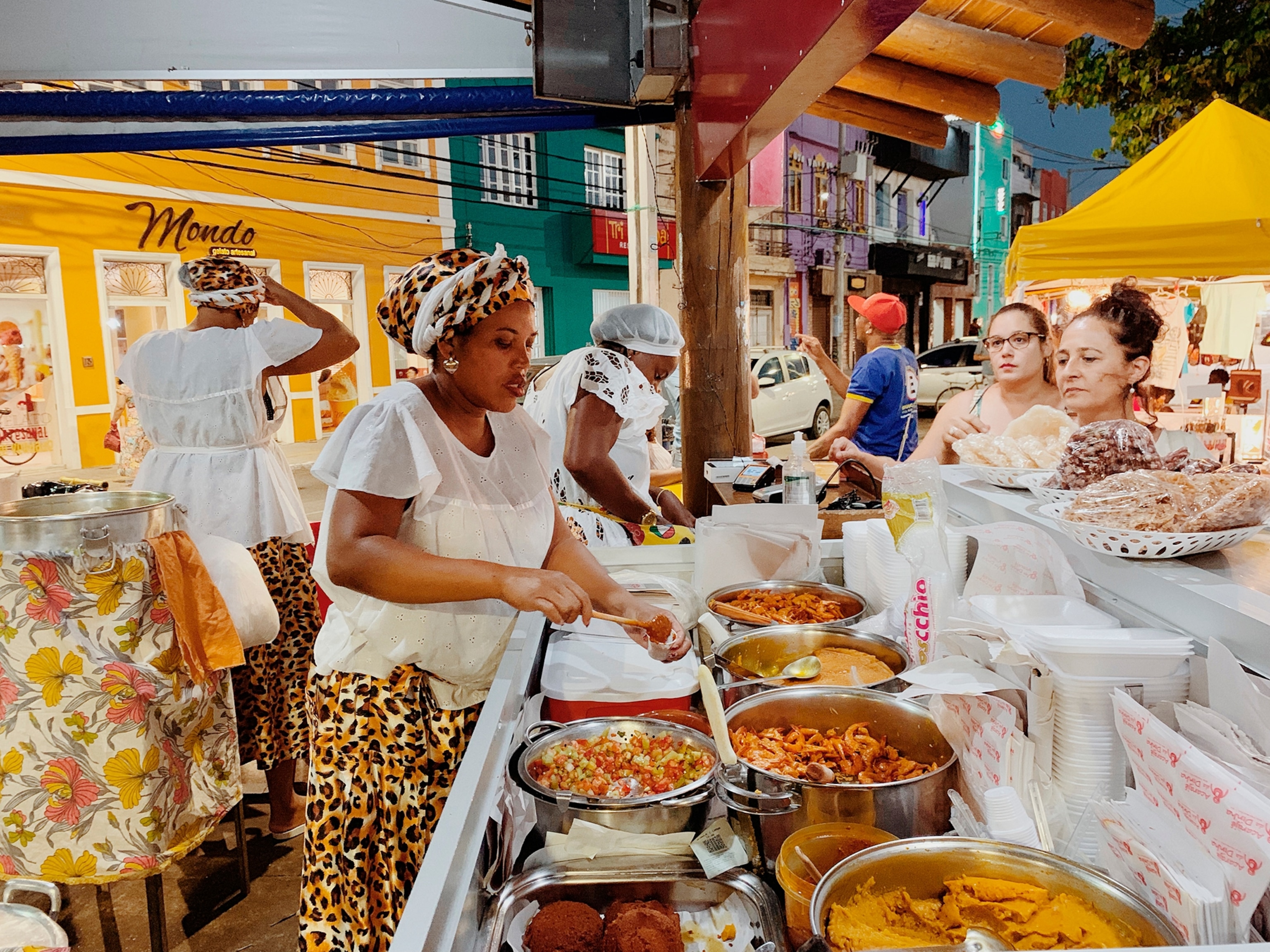 A street food vendor serves prepared dishes from large metal trays at an outdoor market, while customers browse the stand and colorful buildings line the street in the background.