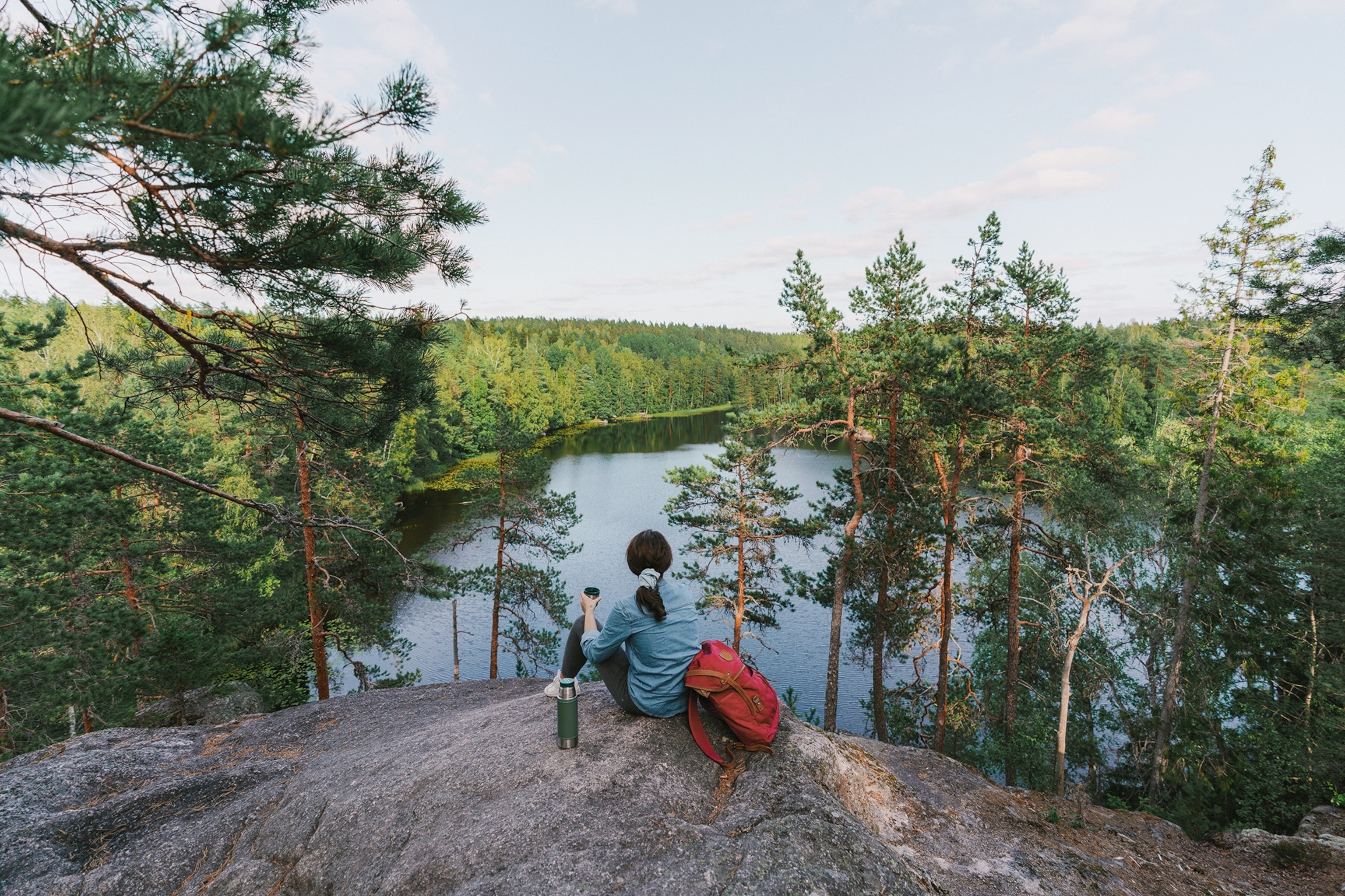 A hiker taking a break atop a rock cliff looking at a lake below with trees around.