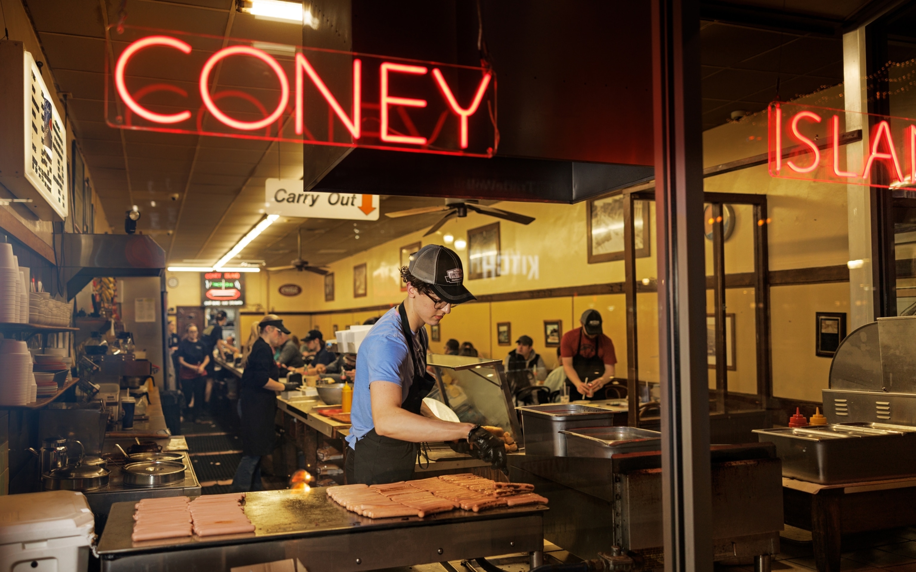 A look through the window at a worker flipping dozens of hot dogs on a grill. A neon sign saying Coney Island hangs on the window.