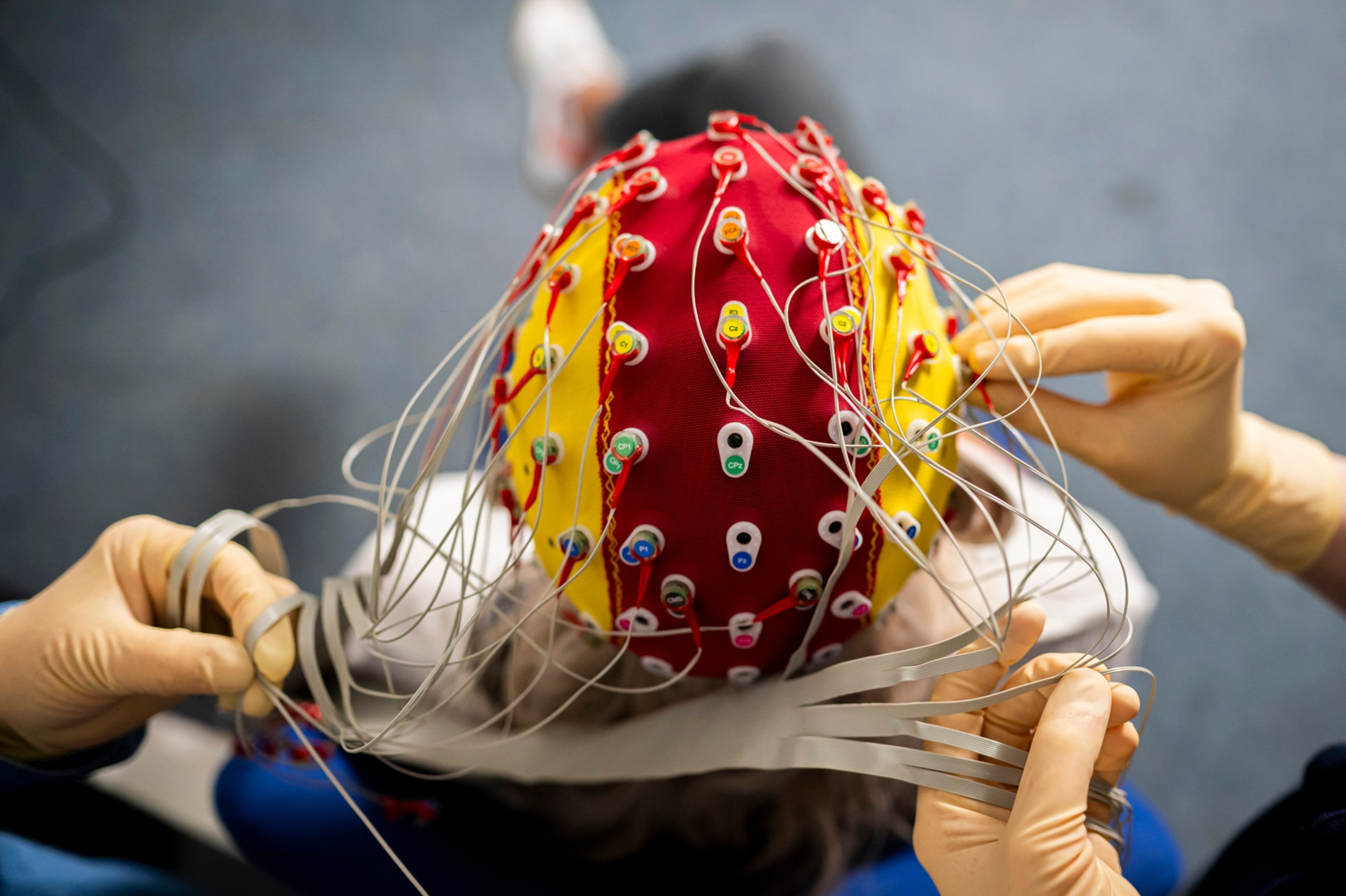 Person wears a yellow and red cap while two people attach electrodes for an EEG.