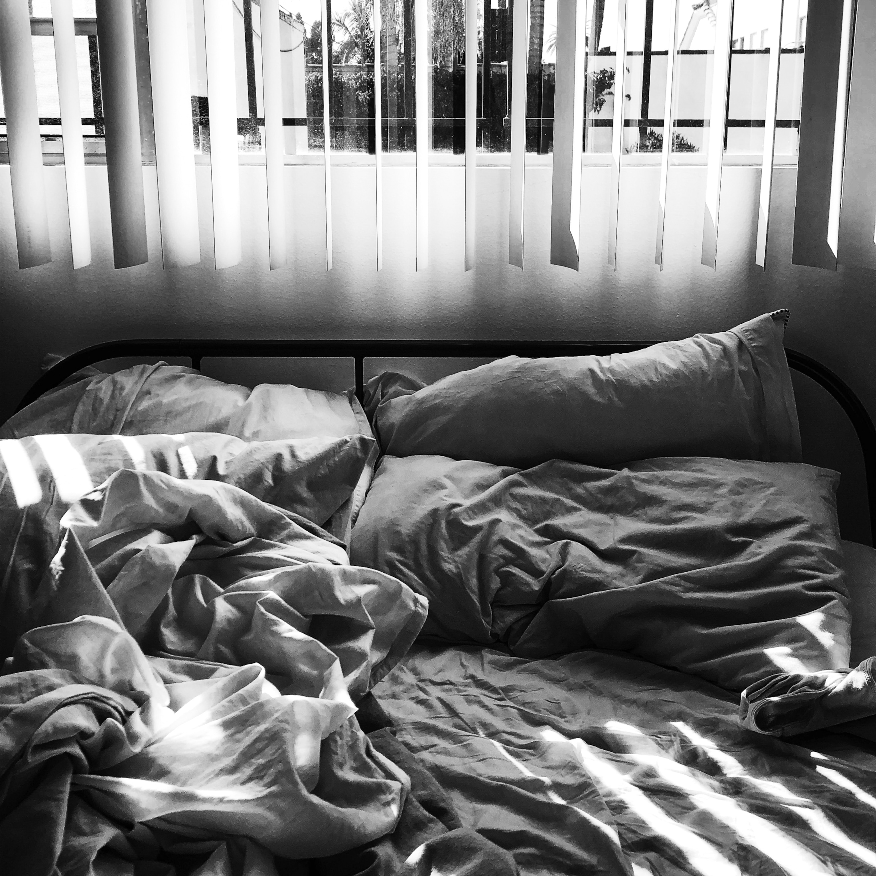 A rumpled bed with white sheets and pillows is lit by sunlight streaming through vertical blinds.