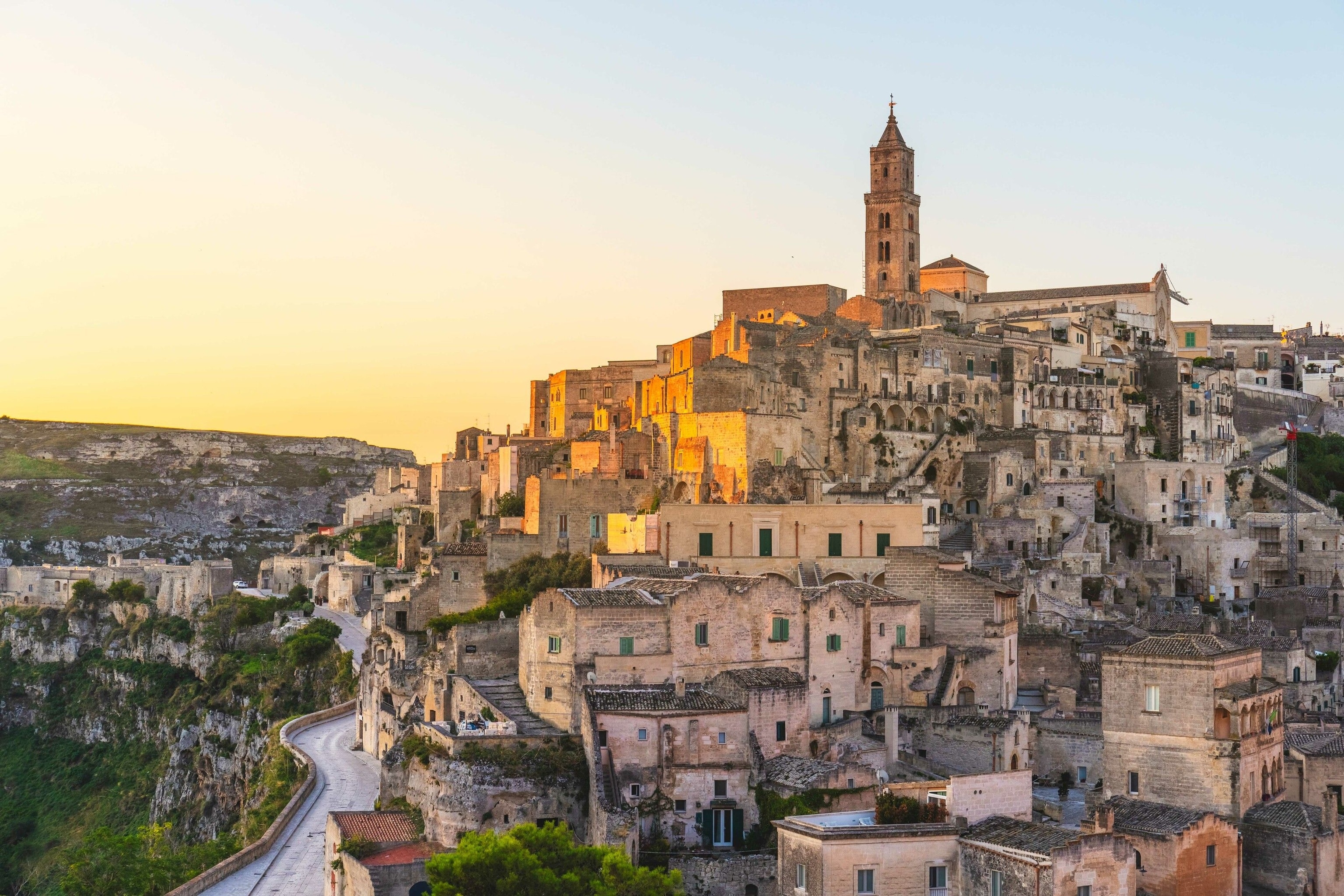 Sunset on the Sassi di Matera. The town is on a hill, and has snaking roads that wind upwards. The buildings look old. They are made of bricks and of stone. A church spire is visible at the top.