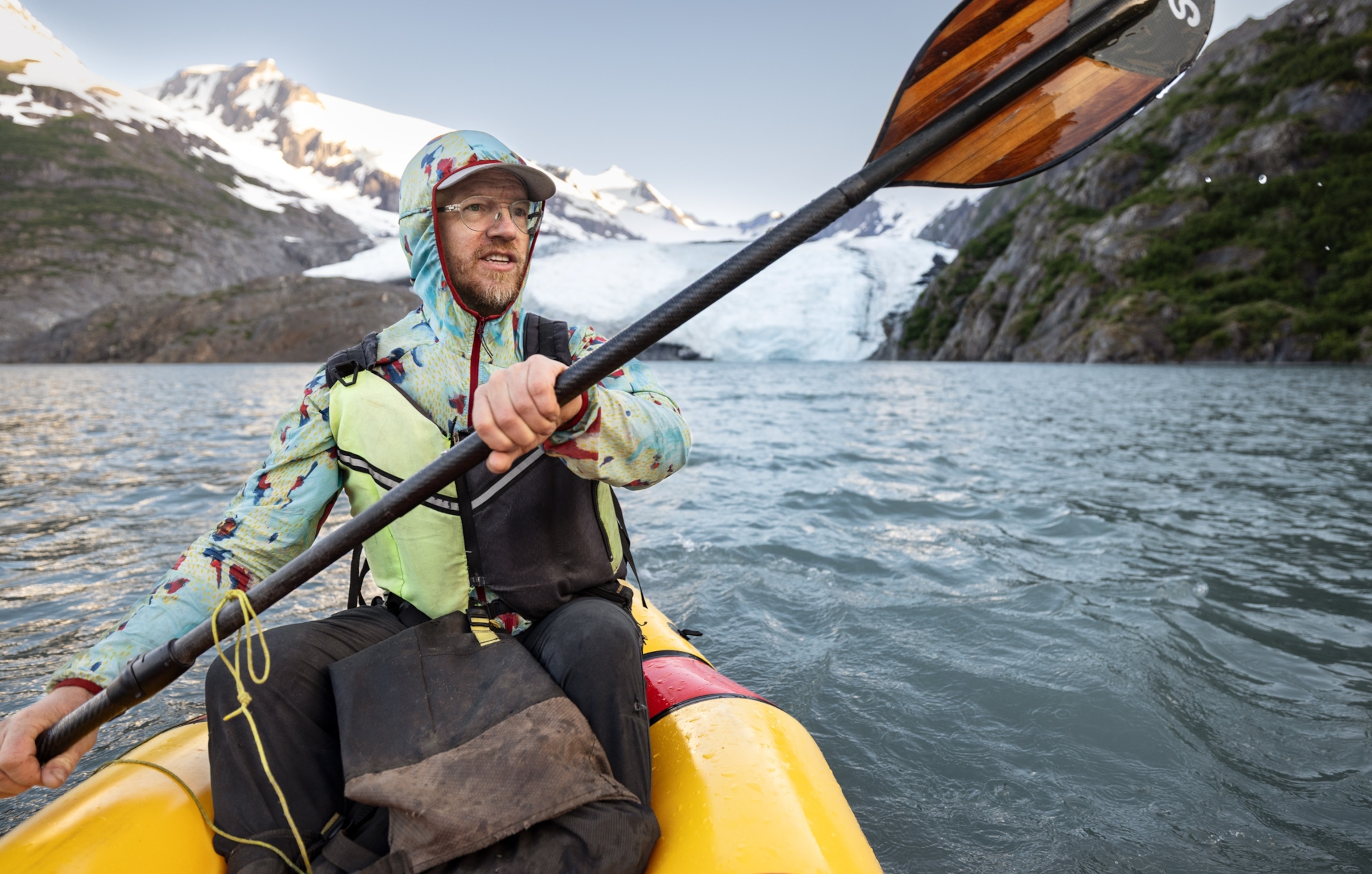 A person paddles a yellow kayak on cold blue water with snowy mountains and a glacier in the background.
