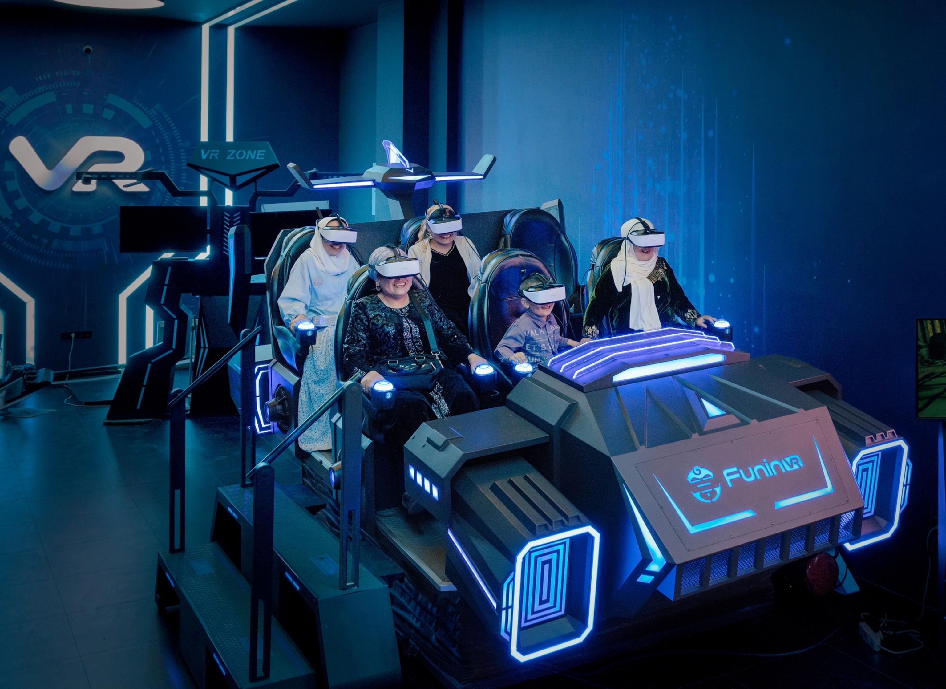 A group of 4 people sit in a futuristic, pretend car as they wear white VR masks that cover their eyes from ear to ear. The car sits in a large blue room with white light strips across the ceiling and wall. A large image of a man wearing the VR mask is projected on the wall next to the car.