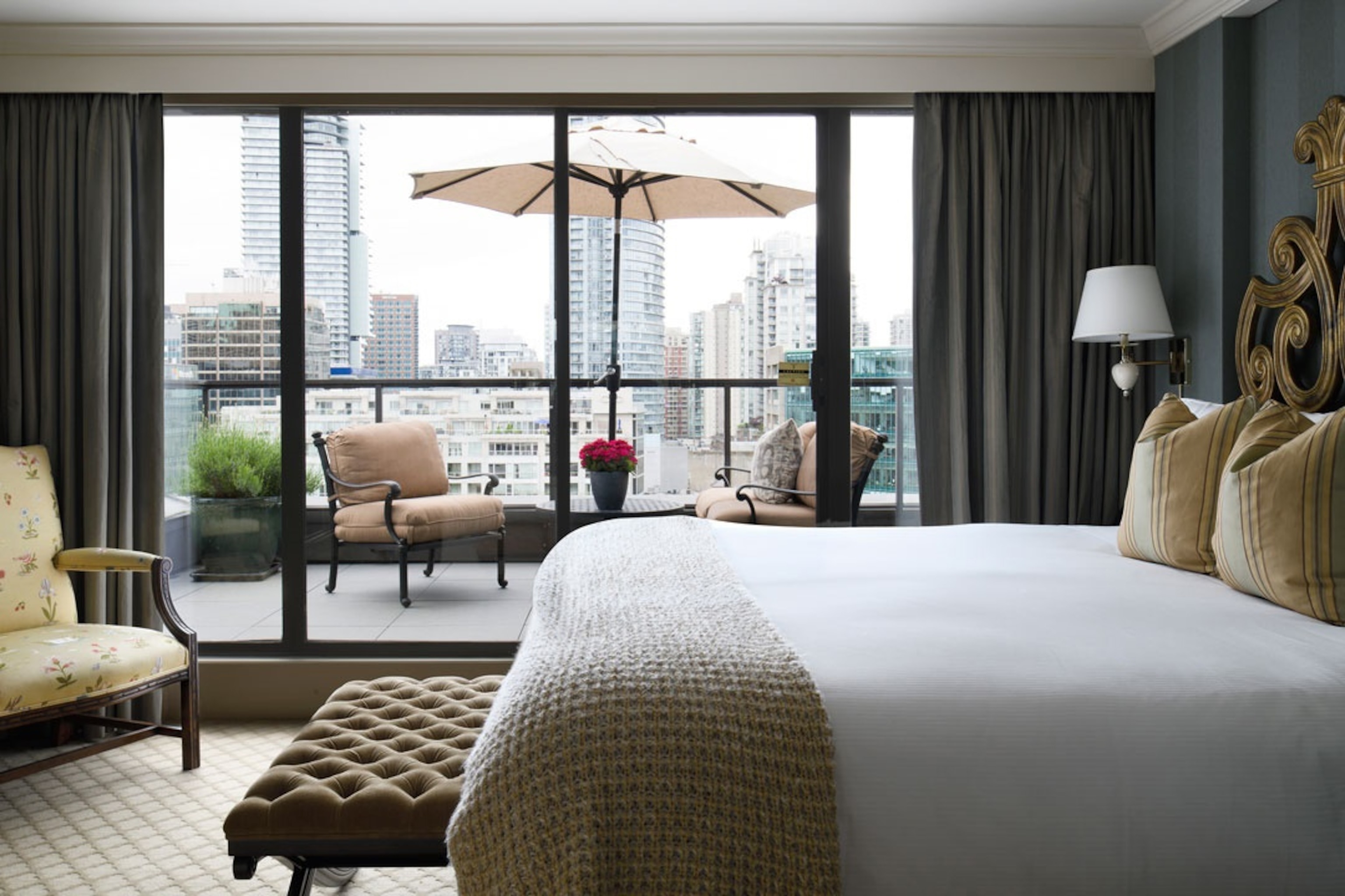 The interior of an elegant hotel bedroom leading out to a balcony with views over the city skyline.
