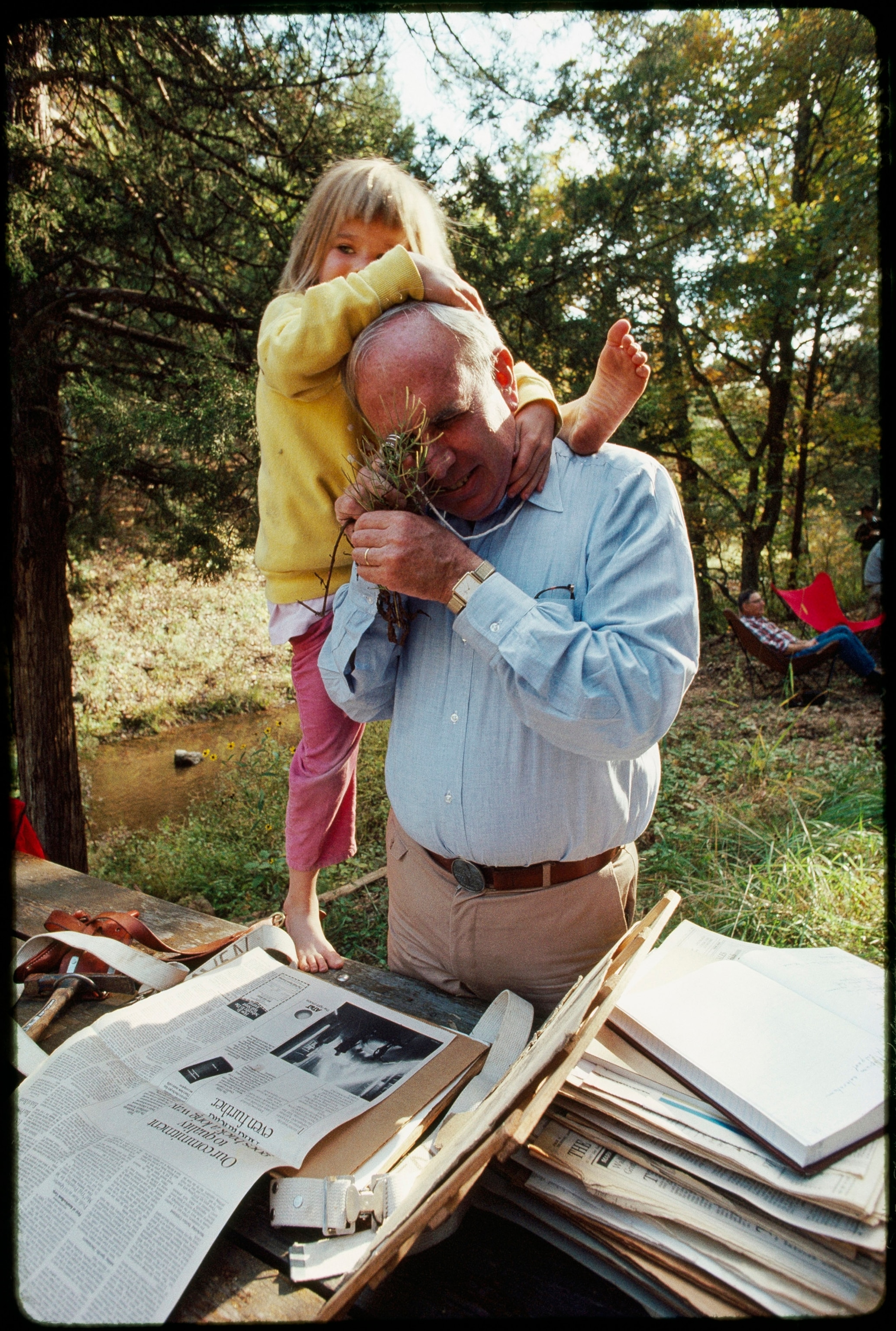 A man in a blue button down shirt peers into a magnifying glass at a leaf while a young girl in pink leggings climbs onto his shoulders.