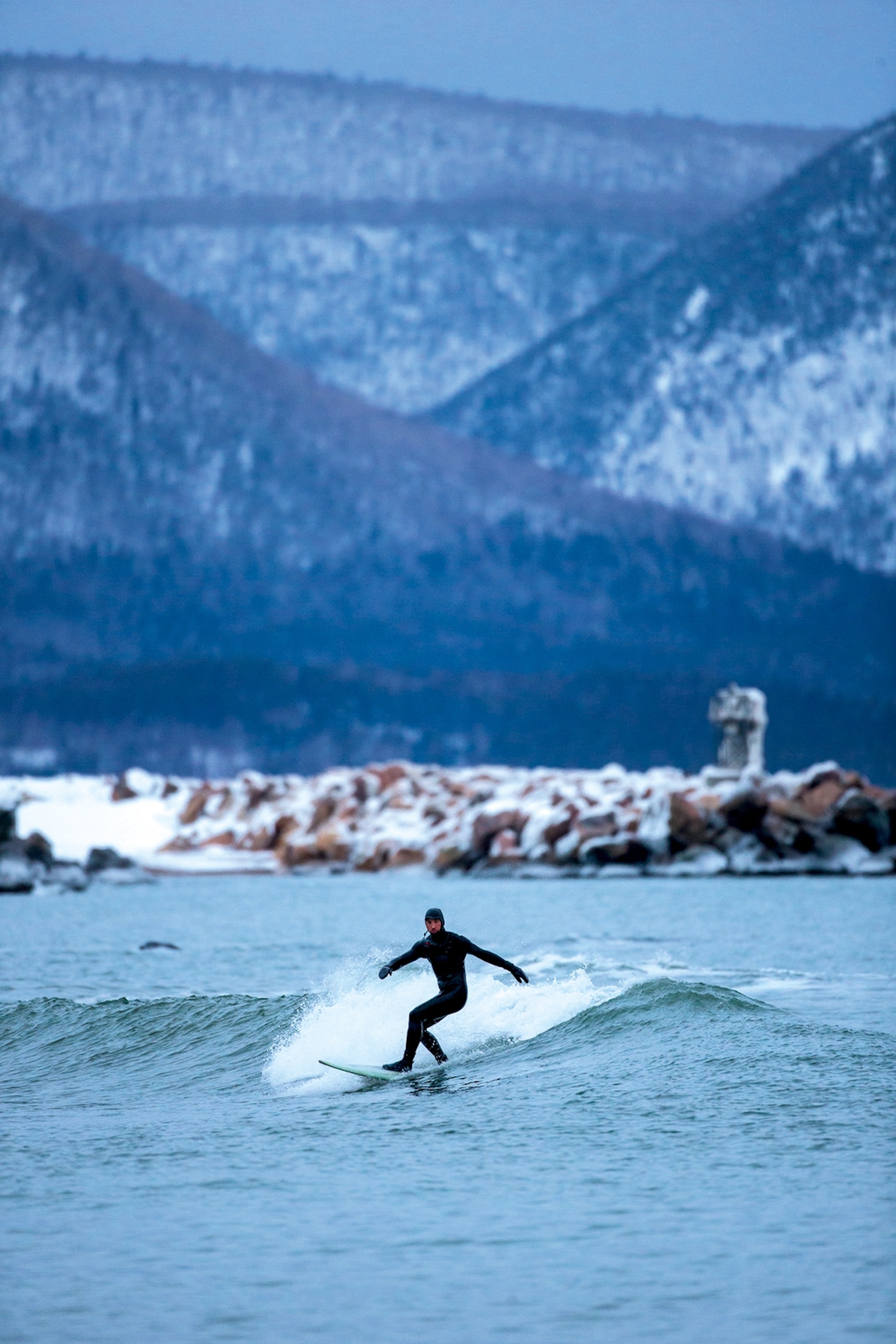 A man in a thick neoprene suit surfing a wave with a snowed-in landscape and mountain in the background.
