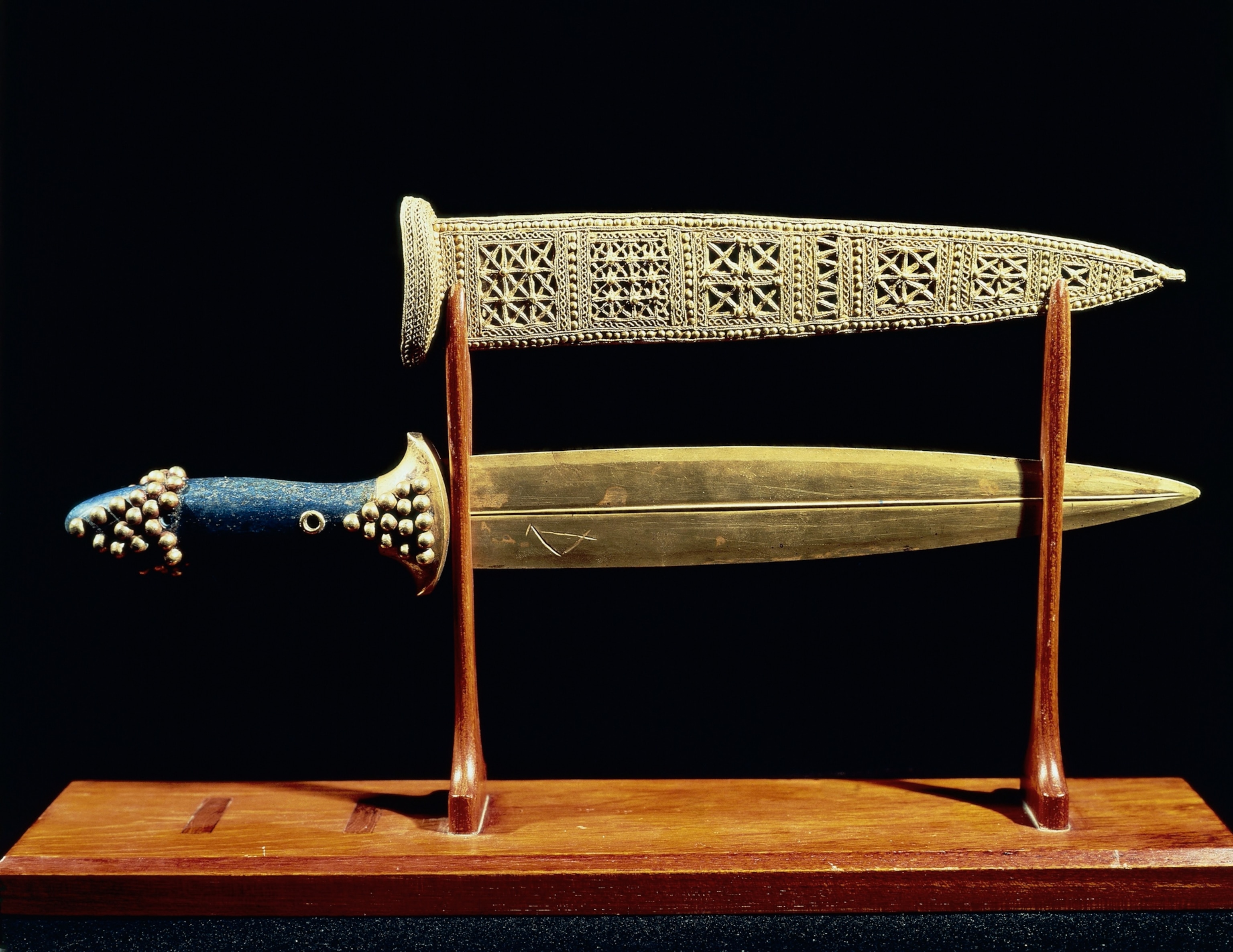 A decorative dagger with an ornate sheath displayed on a wooden stand. The handle is blue with gold accents, and the sheath features intricate geometric patterns