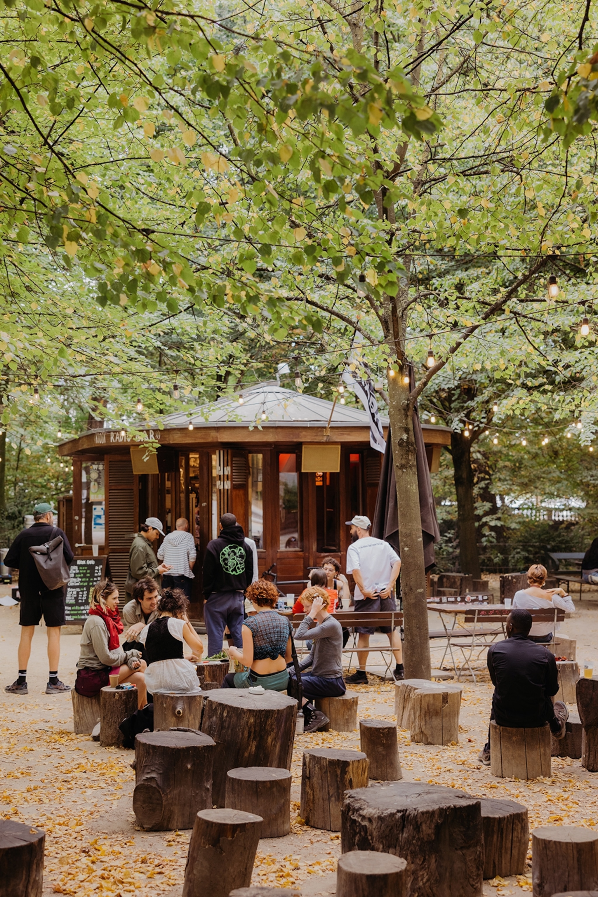 A cosy, open park square with tree trunks as seating opportunities and a small hut with fairy lights and a kiosk menu outside.