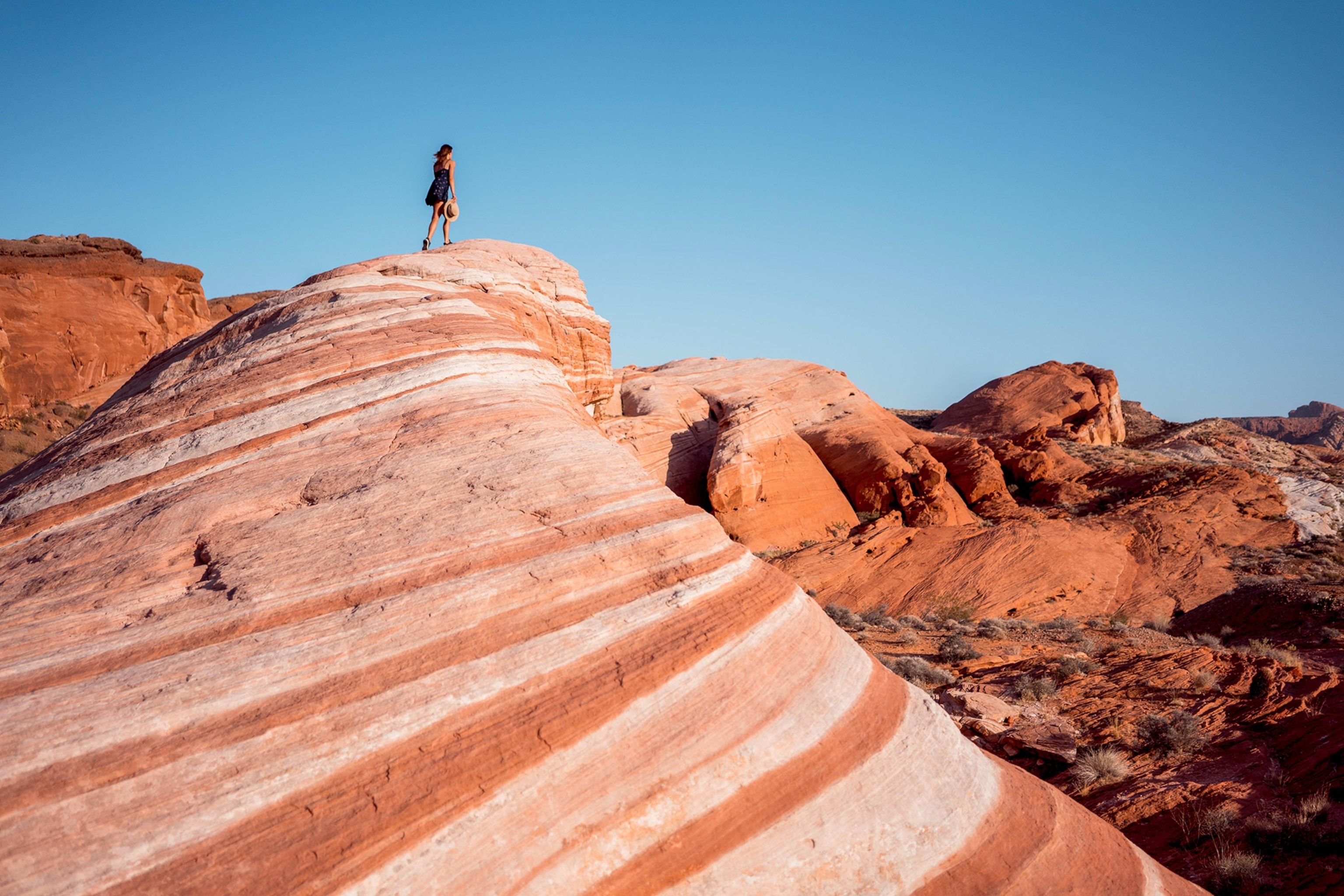A wide shot of red rock formations beneath a blue sky. A woman in the distance stands atop a rocky peak holding a hat in her hand.