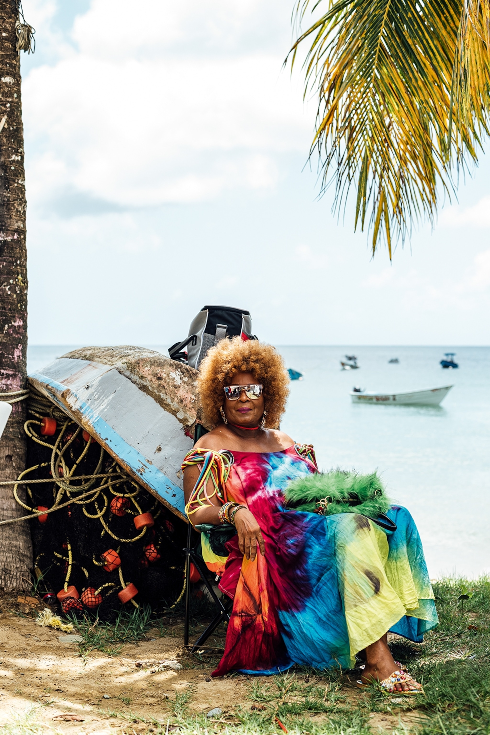 A characterful woman with afro and tie-dye dress sat at the end of a flipped boat by the ocean.