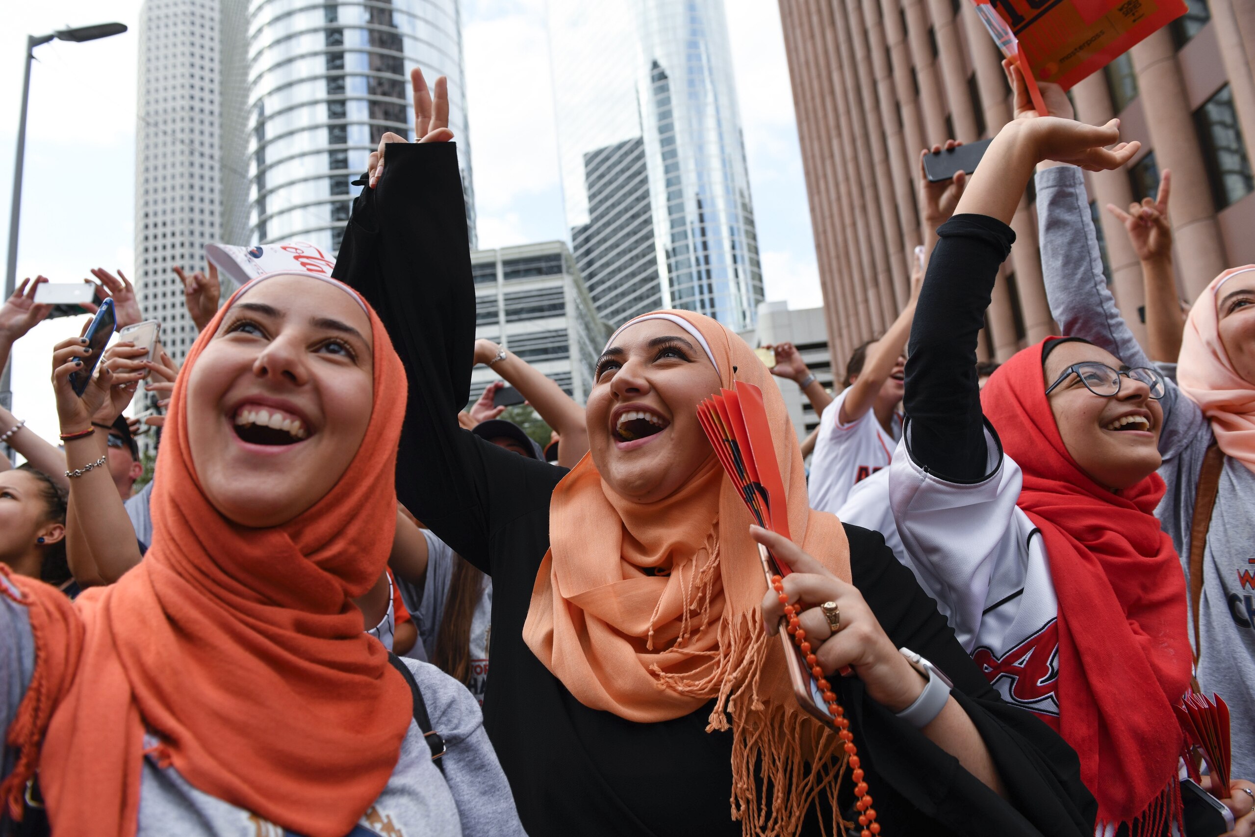 Un grupo de mujeres que visten varios tonos de hijabs anaranjados, sonríen brillantemente y saludan.