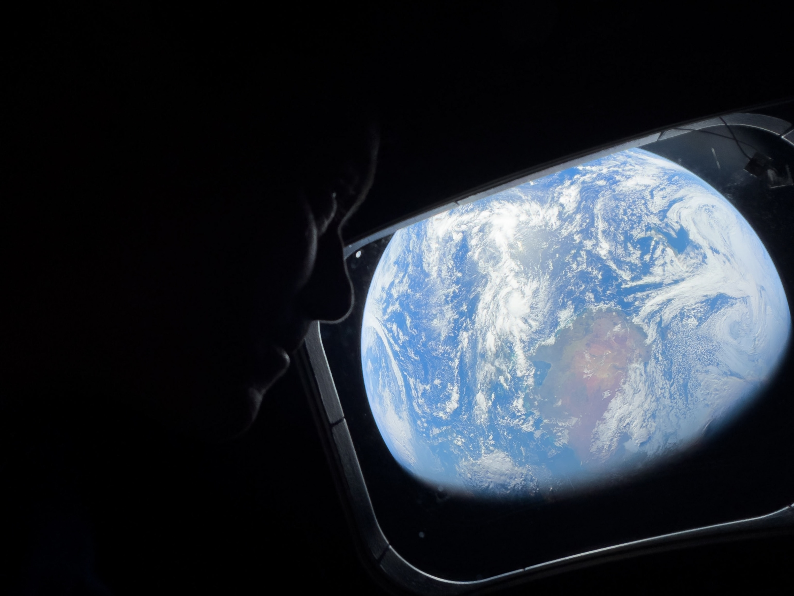 A person gazes out a spacecraft window at Earth, which appears vibrant blue with swirling white clouds against the dark expanse of space.
