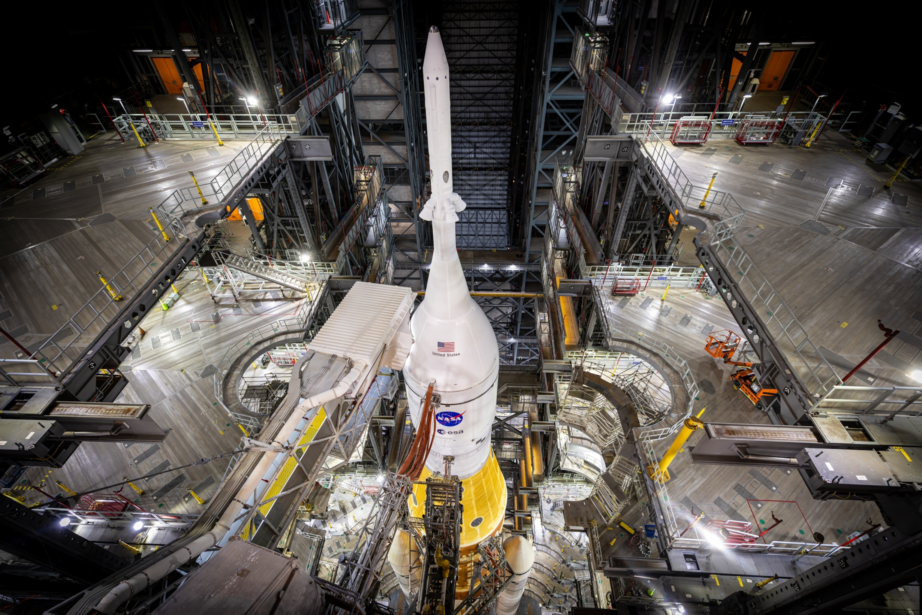 A large rocket stands vertically inside a spacious industrial hangar. Bright lights illuminate its white exterior and complex surrounding scaffolding.
