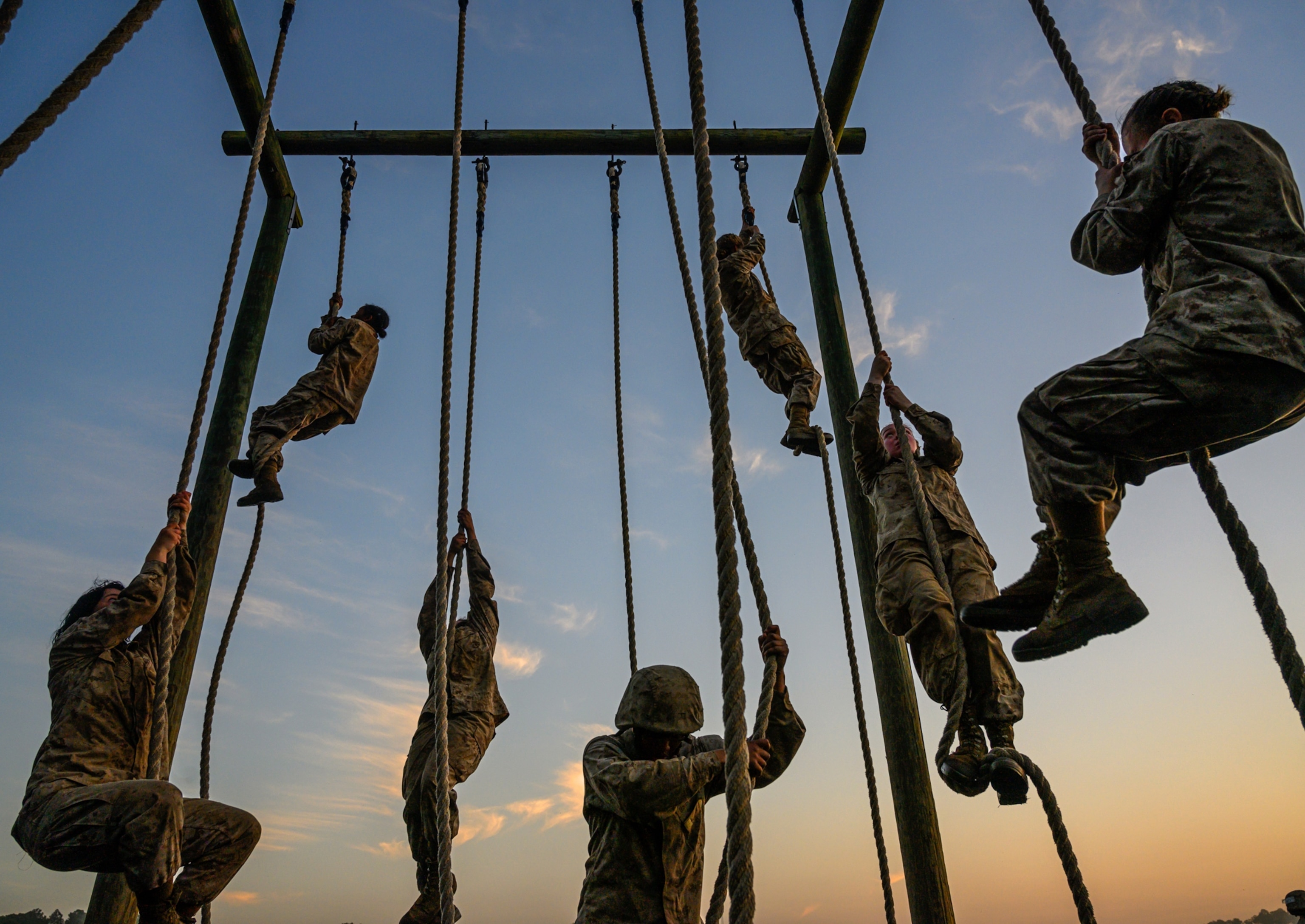 Soldiers in camouflage uniforms climb ropes at an outdoor training course during sunset, silhouetted against a gradient sky