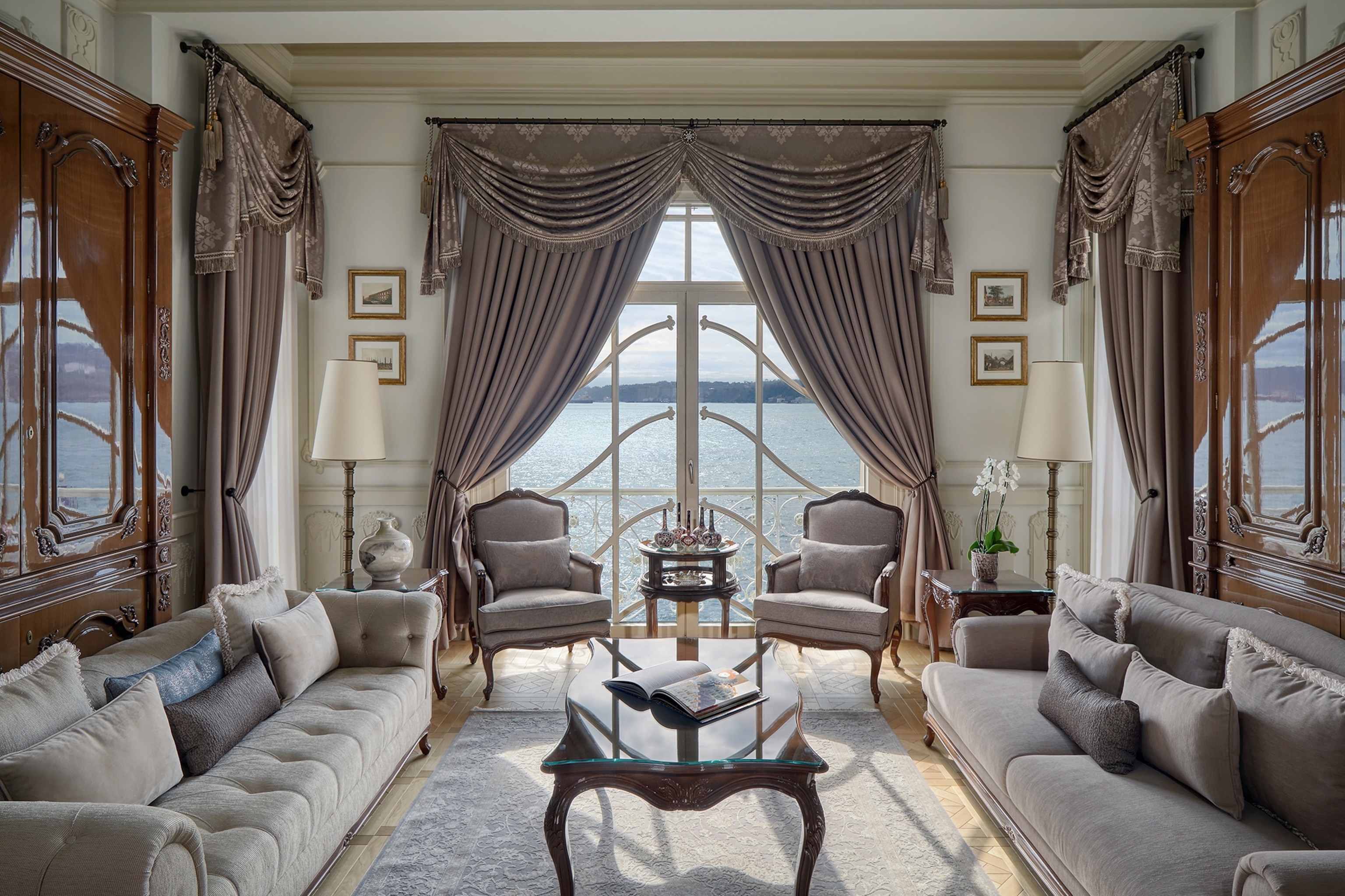 A grand living room area with luxurious chairs and a library on either side; a central window shows the ocean outside.
