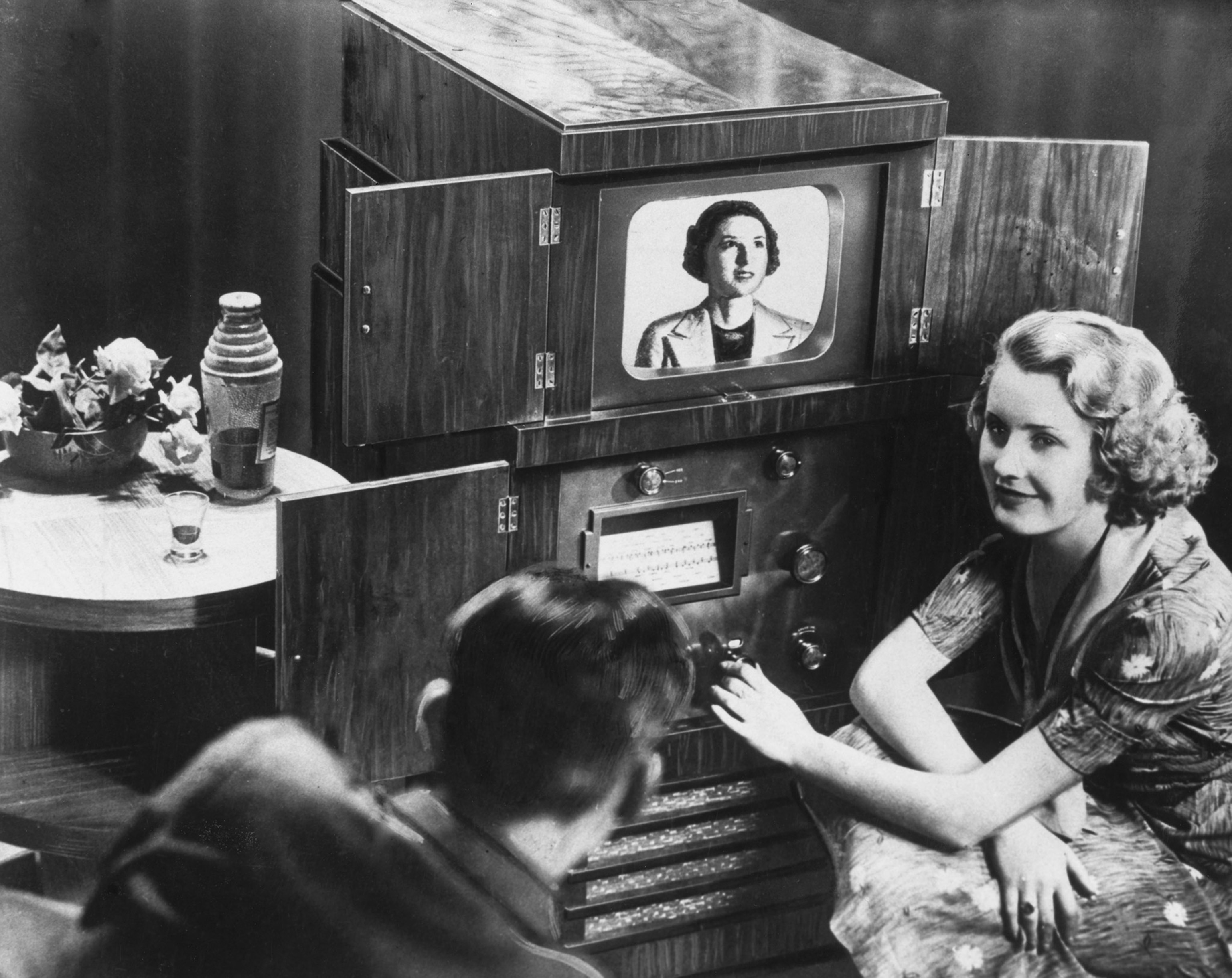 A black and white historical photo, a woman kneels next to an early television model, a man looks on in foreground