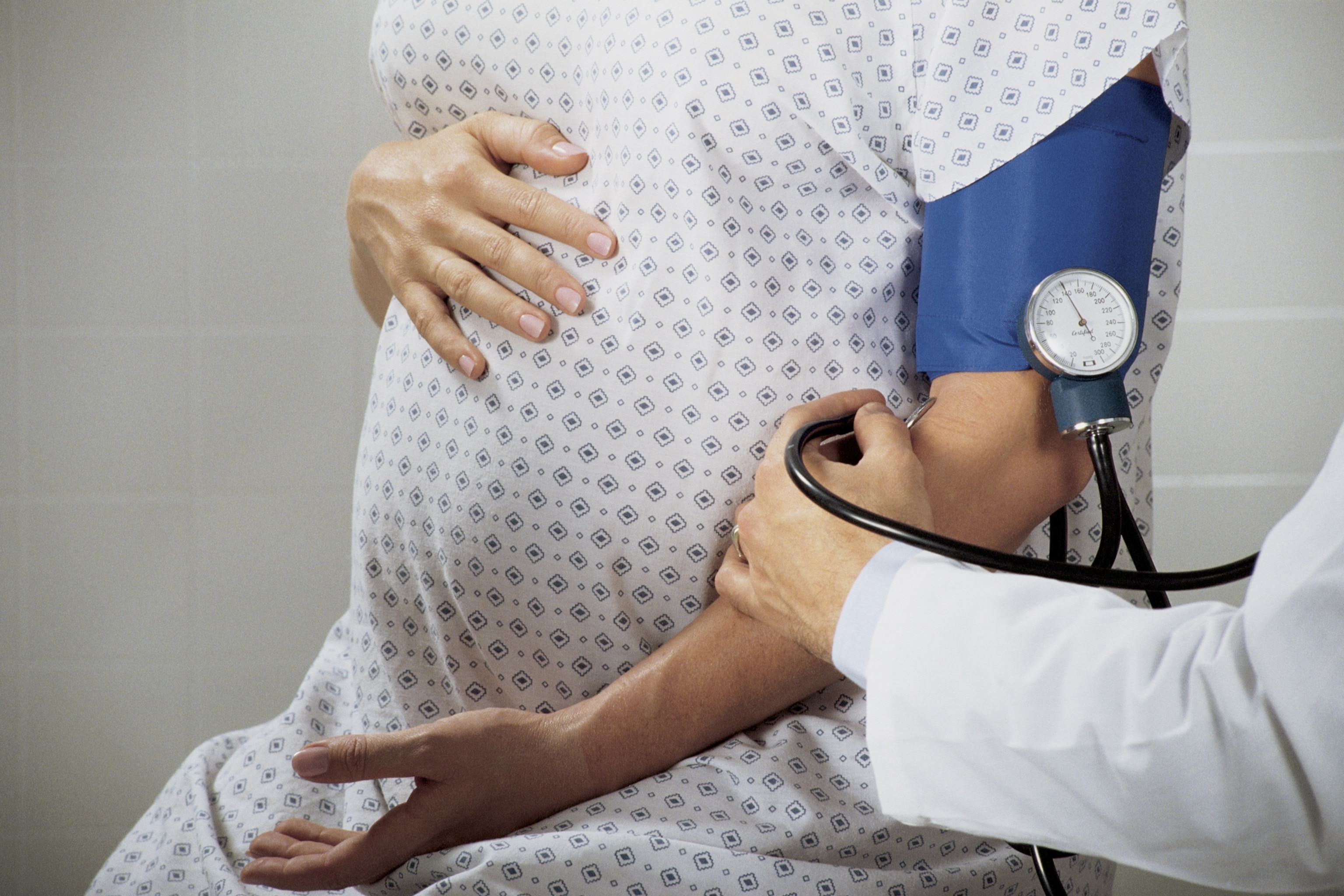A healthcare professional in a white coat uses a stethoscope to measure the blood pressure of a pregnant woman wearing a patterned hospital gown.