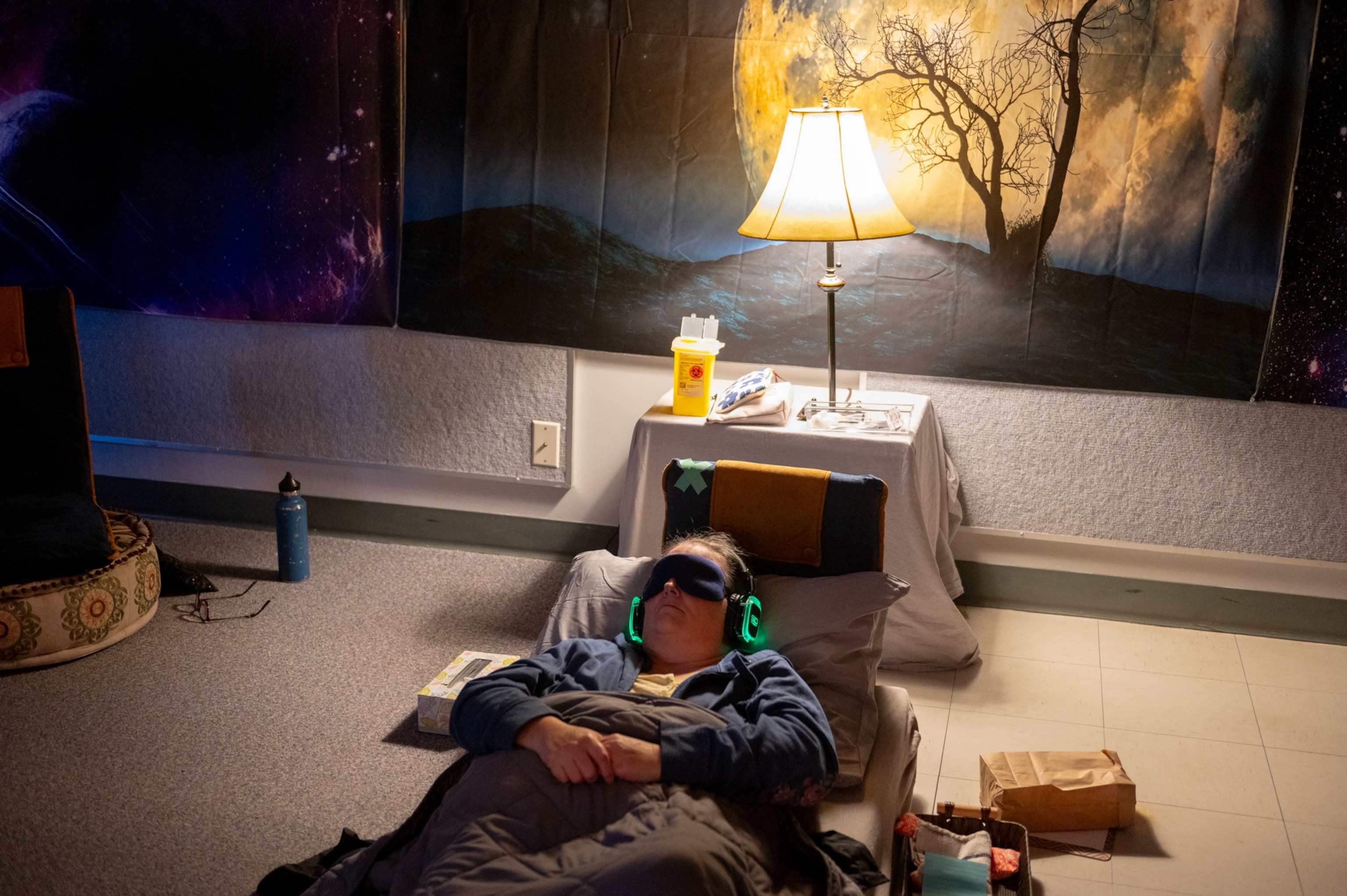 A woman lies on a cot, wearing headphones and a sleep mask, in a dimly lit room with a glowing lamp. The walls feature a cosmic and moonlit mural.