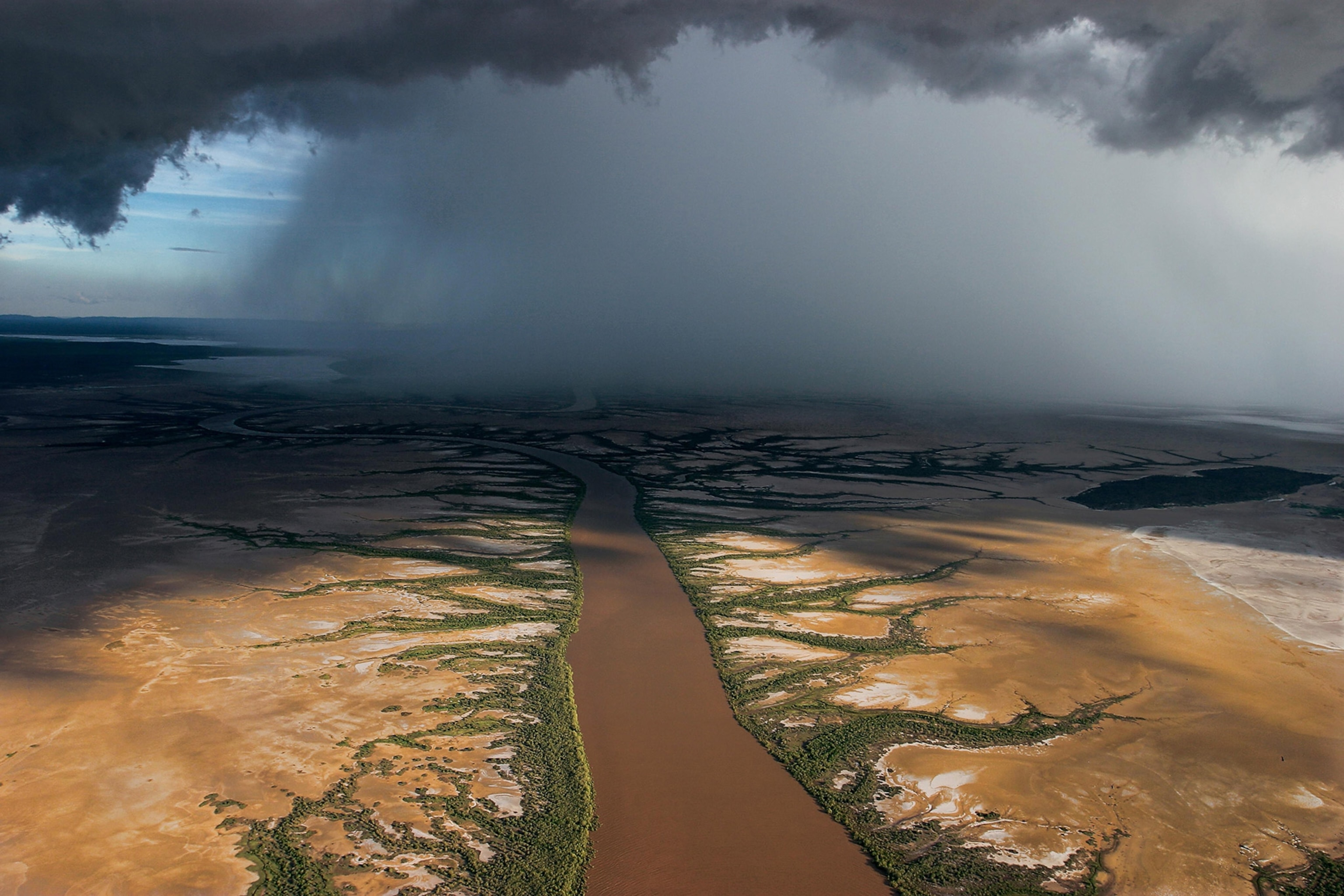 A brown river stretches out toward the horizon with a dark rain cloud dropping heavy rain in the distance