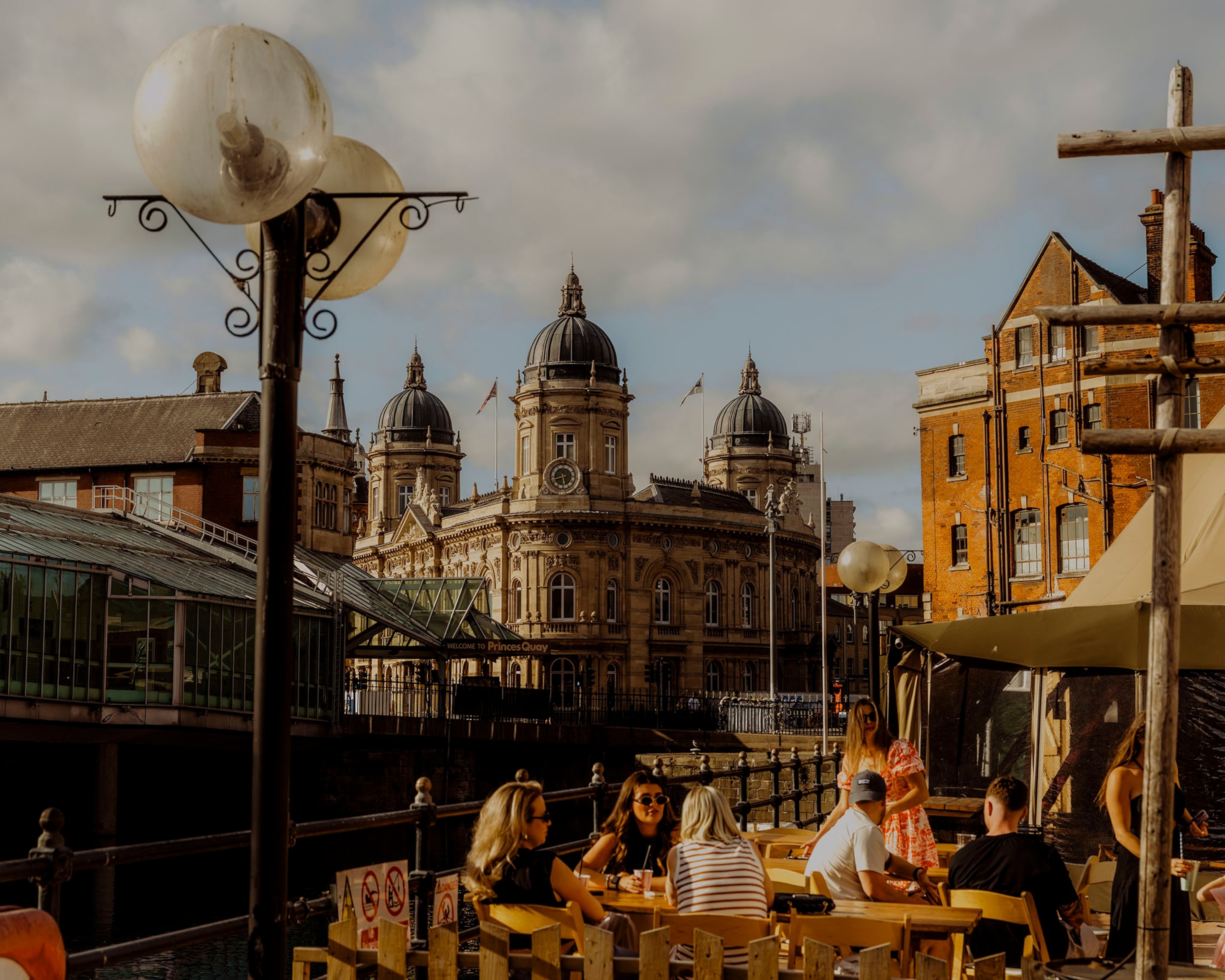Hull, United Kingdom — July 2025: Scenes from Hull’s maritime heritage and community, showing the exterior of the Hull Maritime Museum across Prince’s Dock, with people socialising outside a local bar in the foreground.