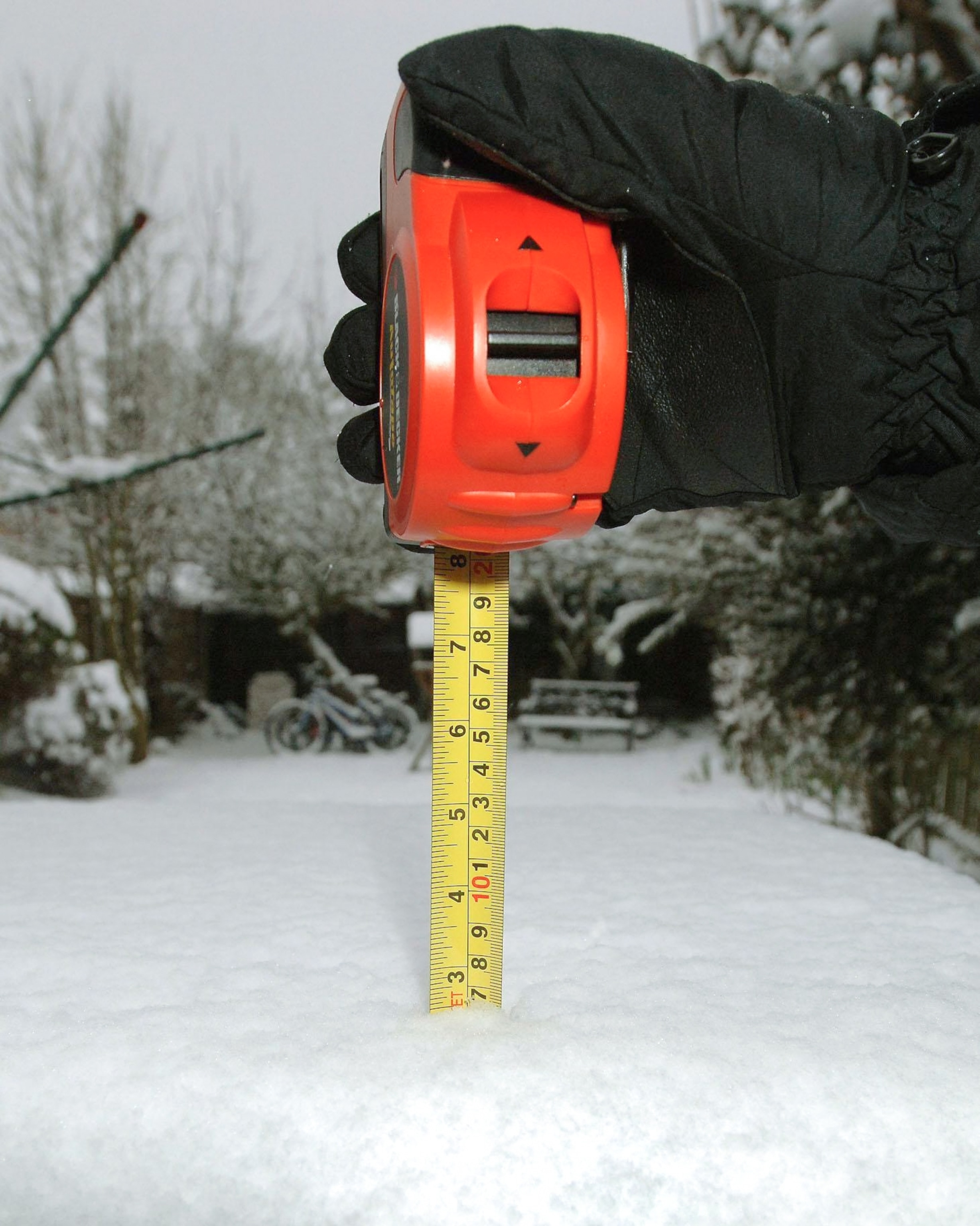 A gloved hand holds a red measuring tape, measuring snow depth on a surface in a snowy yard.