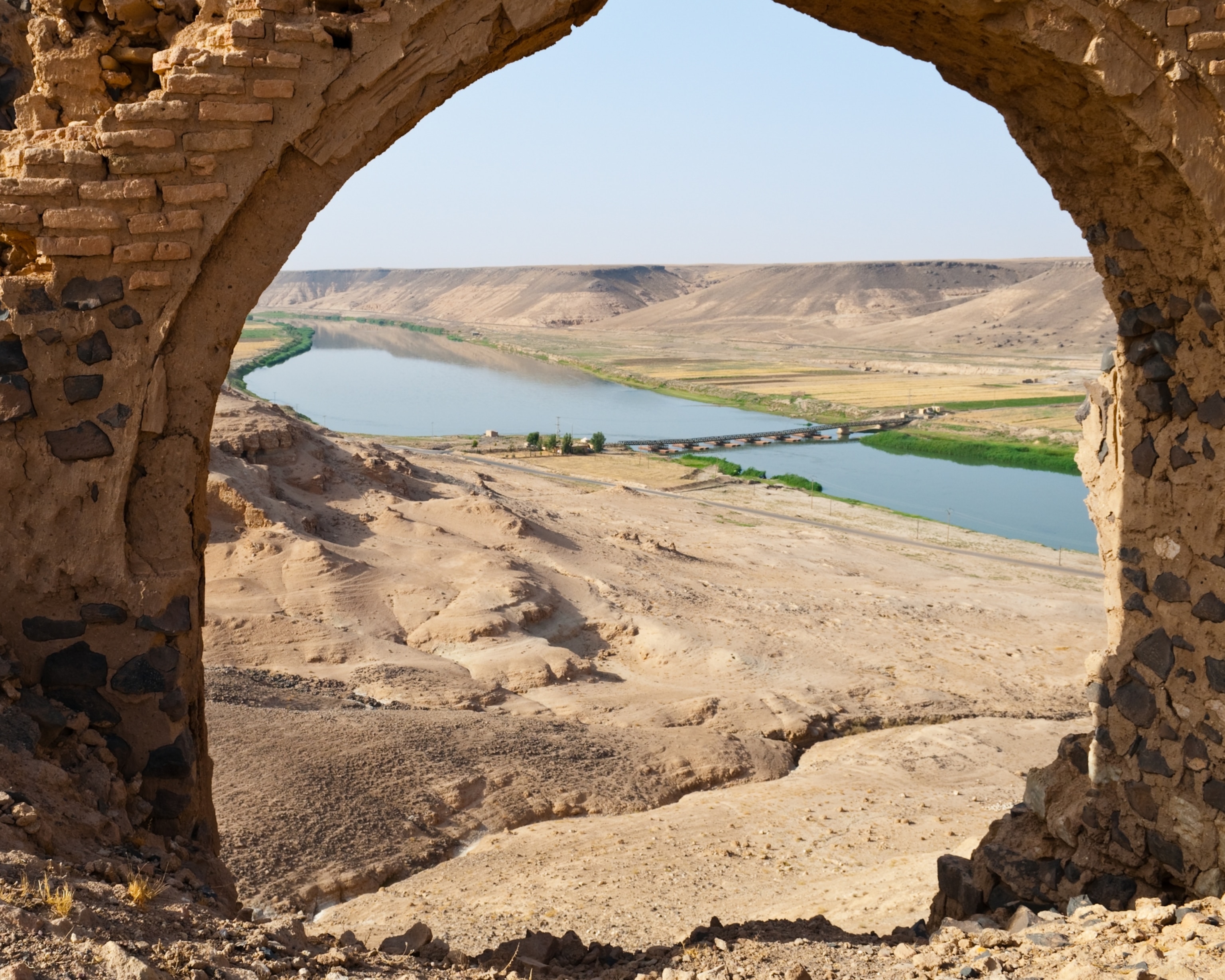 View through a stone archway of a tranquil river winding through a vast desert landscape, with sparse greenery and a bridge