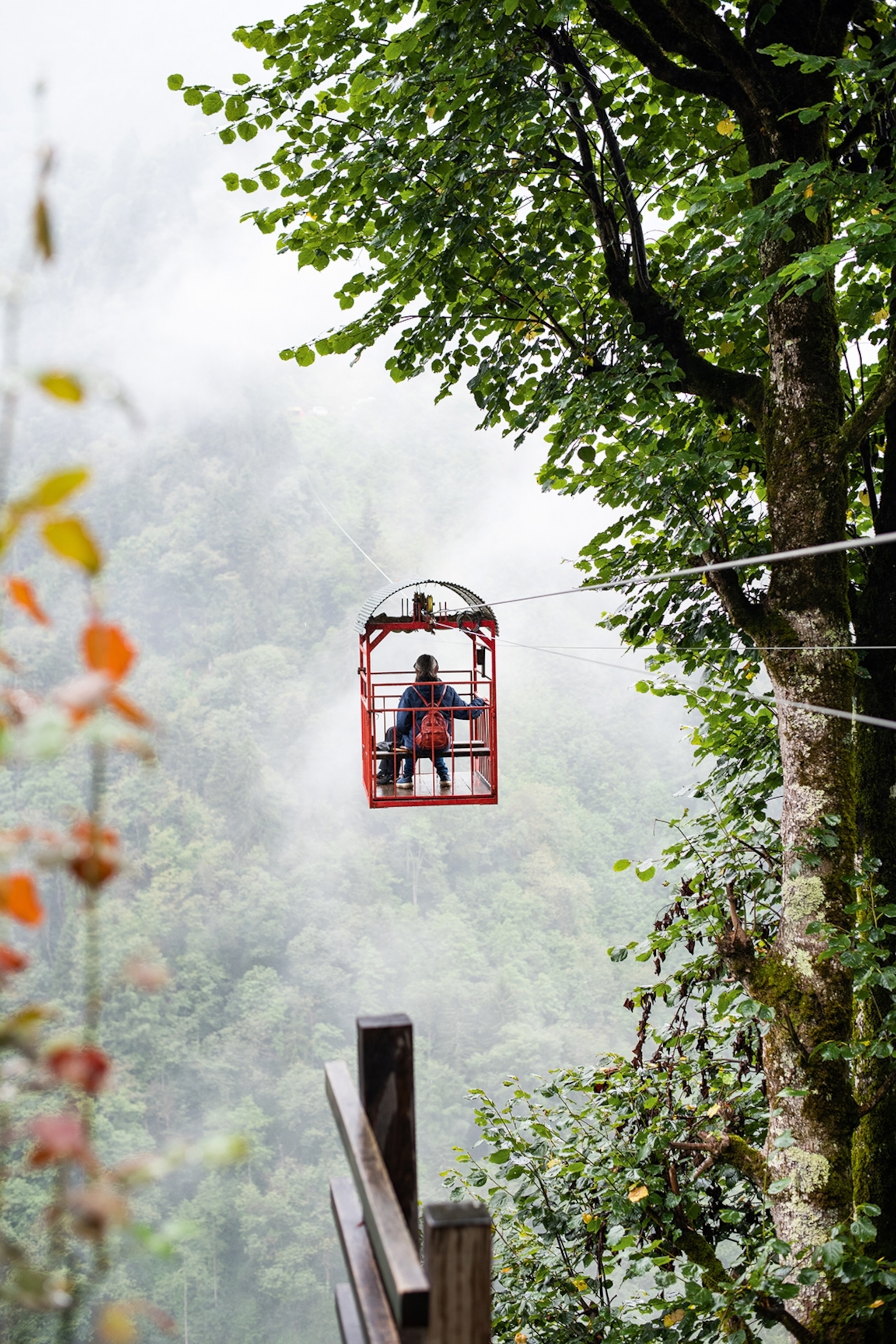 A simple cable car with one person riding down over a mountain gorge with thick forests and fog hanging in the clouds.