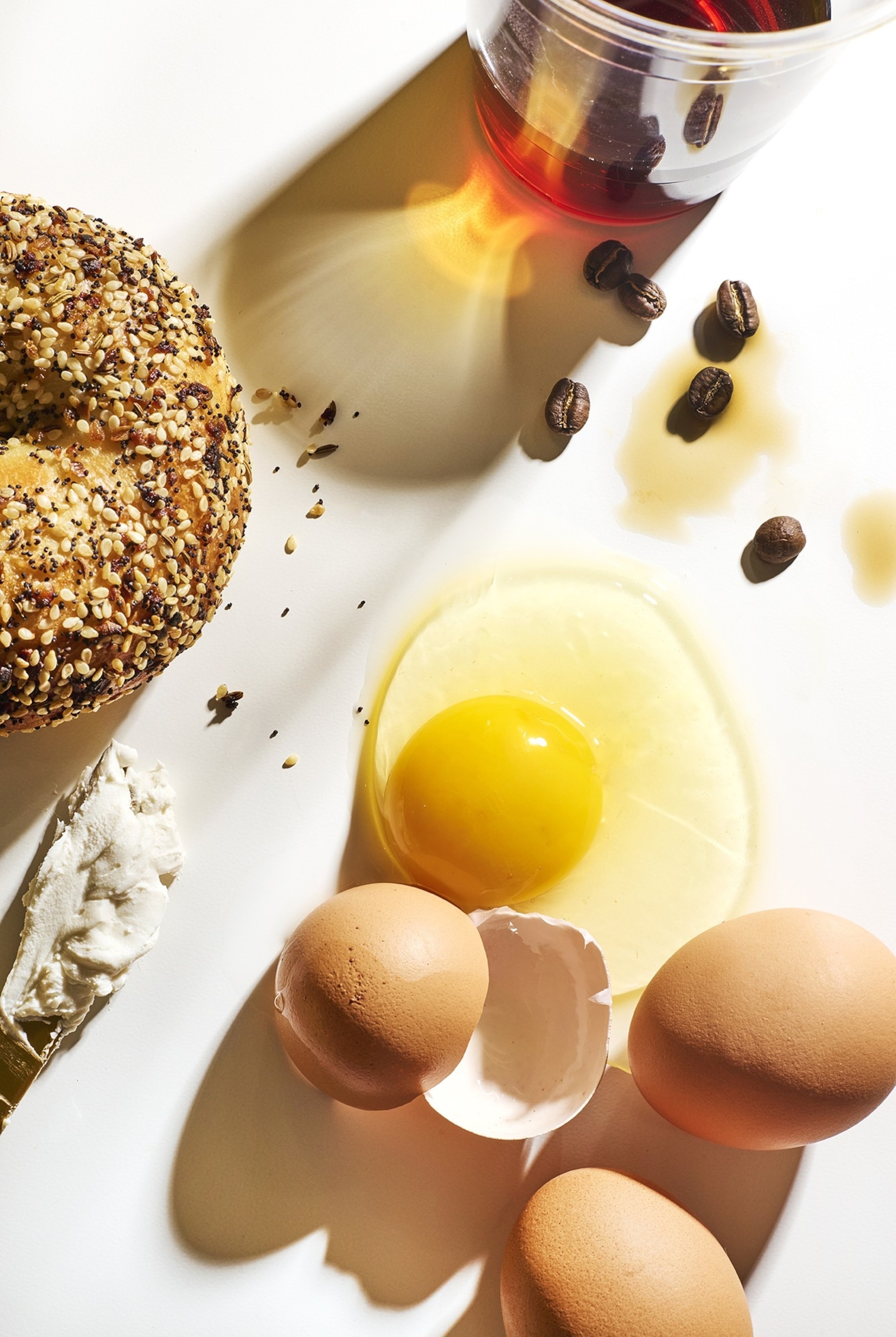 overhead view of cracked eggs, an everything bagel, and coffee on a white background