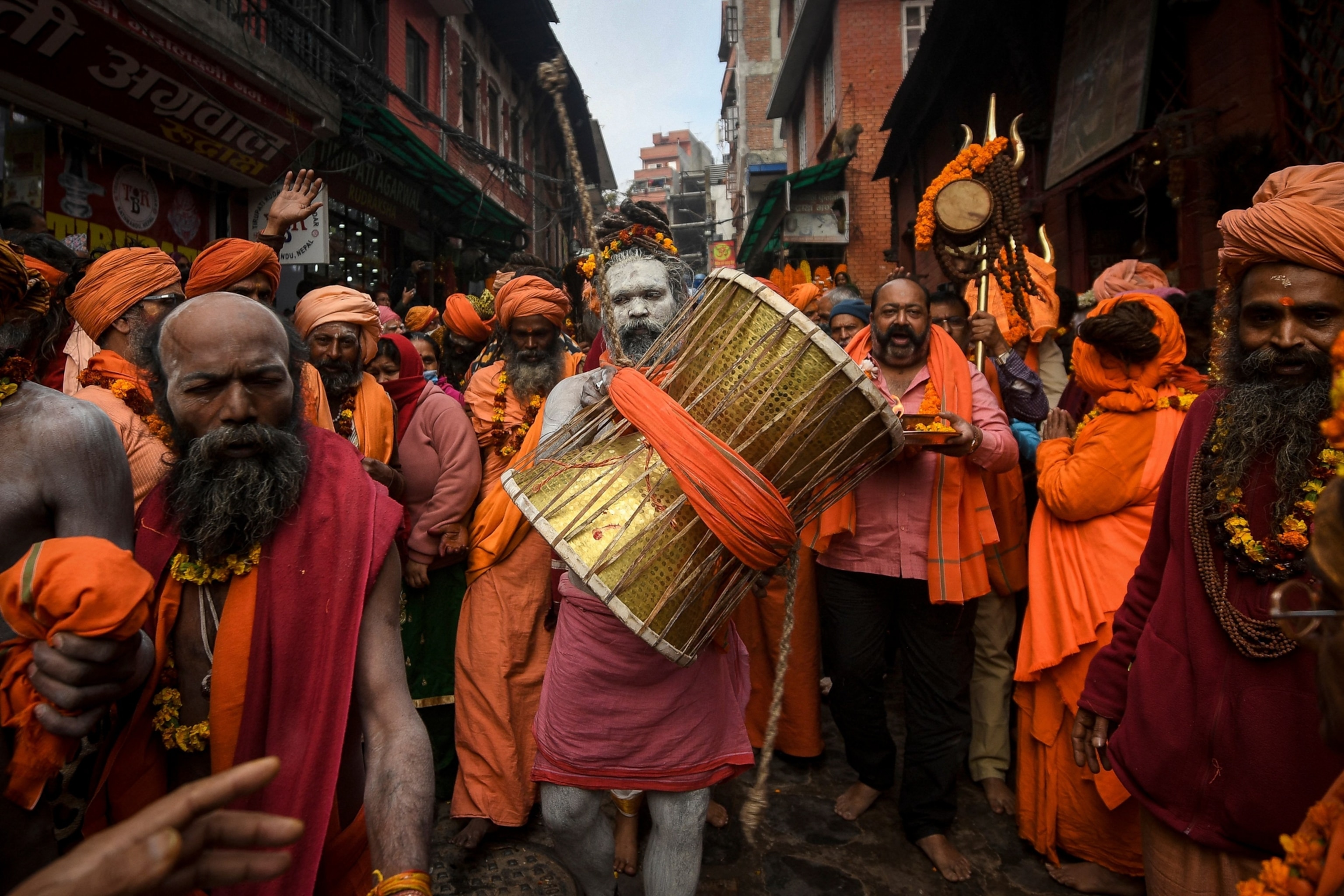 A vibrant festival scene features numerous people in bright orange clothing and headwear. A central figure with white body paint plays a large drum, while others around him joyfully participate. The narrow street is bustling and surrounded by traditional buildings.
