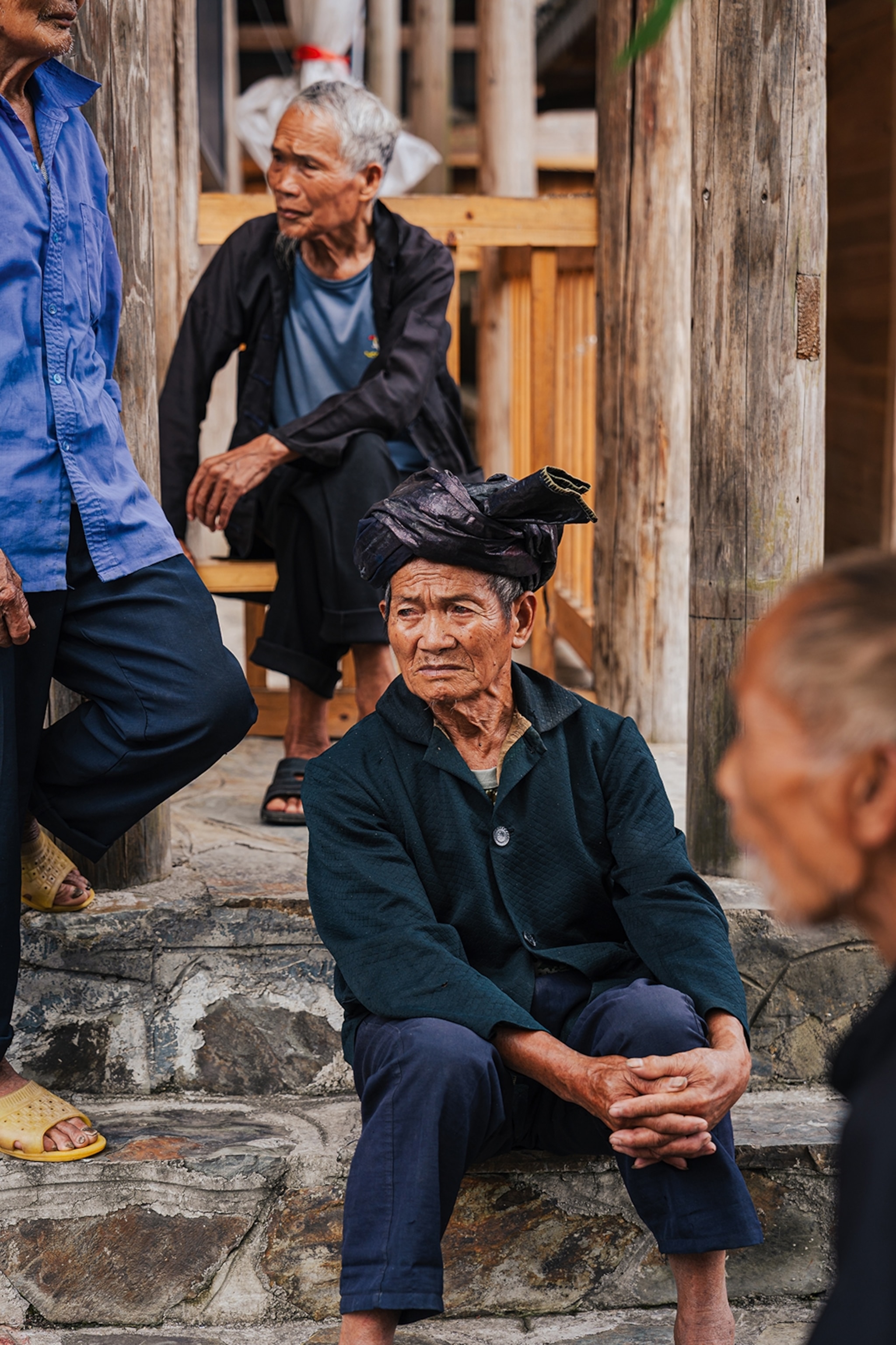 An elderly Chinese man with a simple fabric turban sitting on the stone steps of his house, looking intro the distance with a concerned expression.