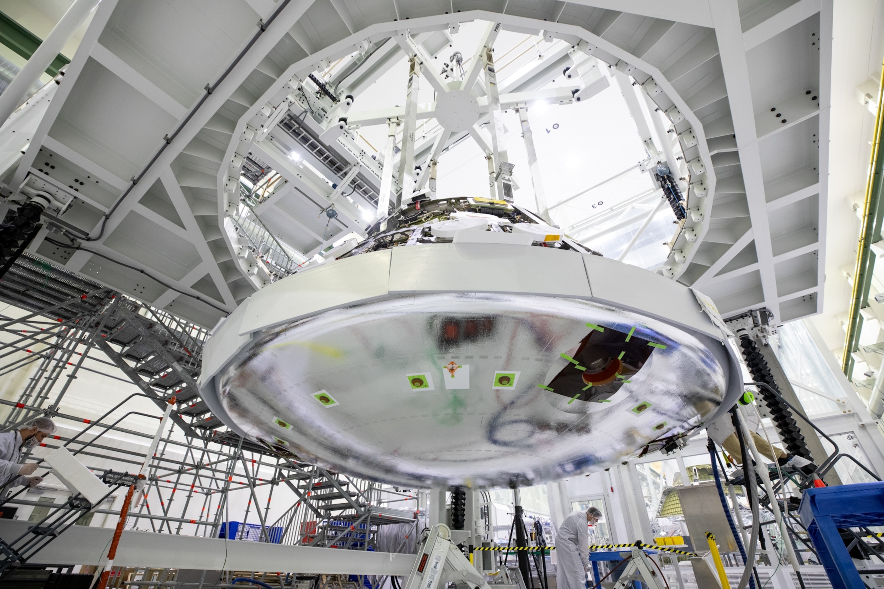 Large spacecraft component being assembled in a high-tech facility. Technicians in protective gear work amidst metal scaffolding