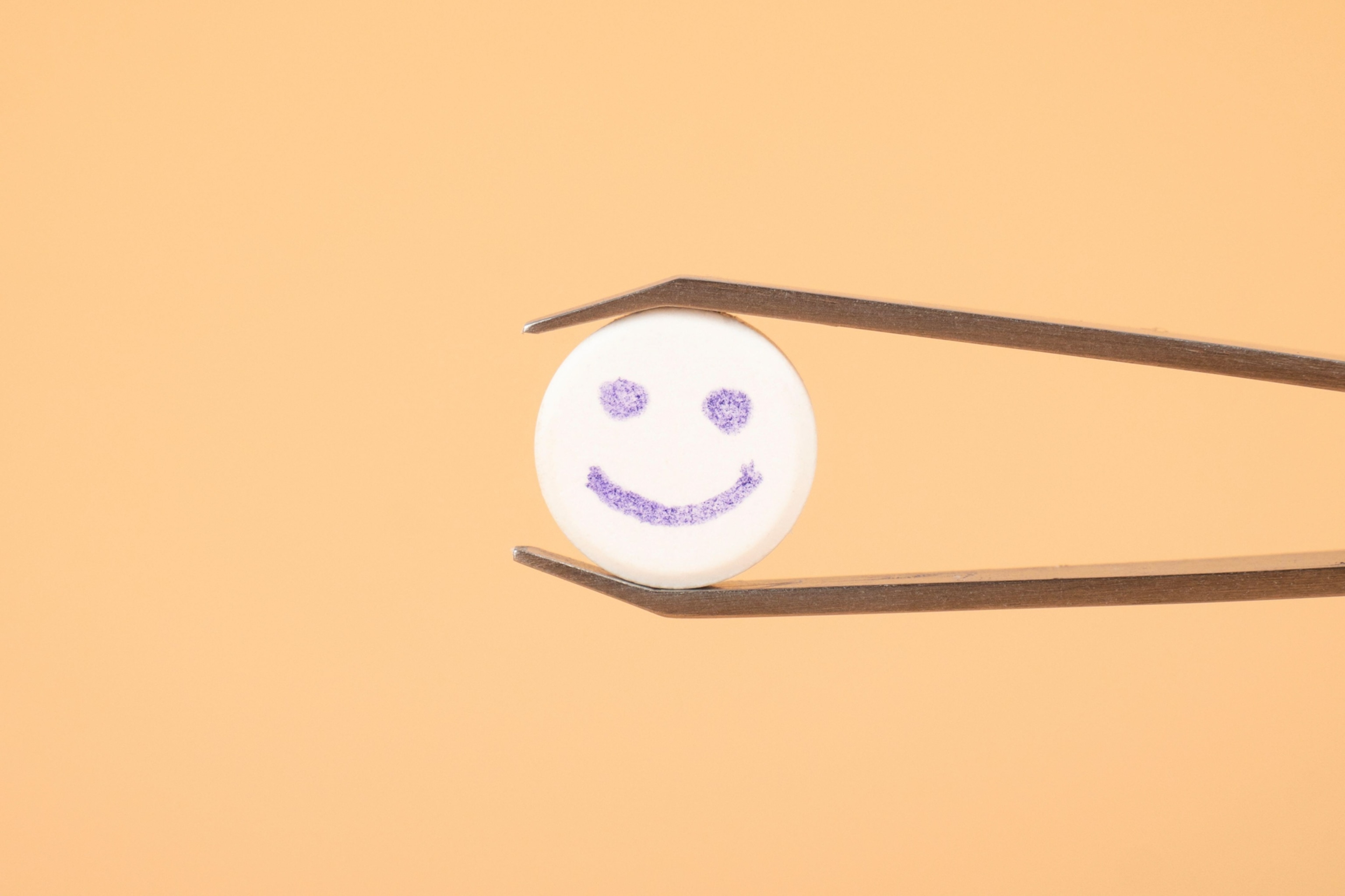 A white tablet with a smiley face drawn on it, held by a pair of metal tweezers