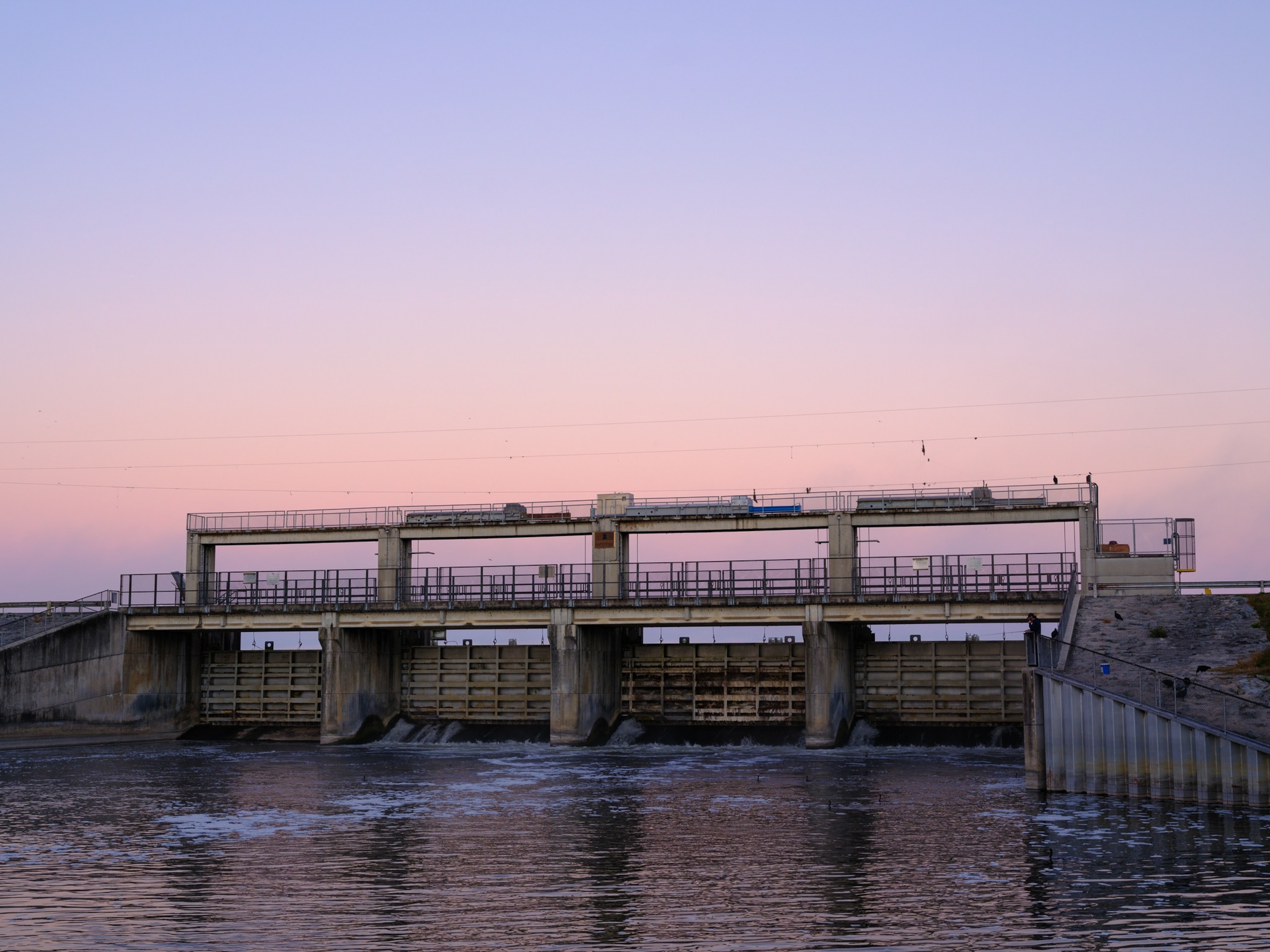 A dam stretches across a calm river under a soft pink and blue sunset sky.