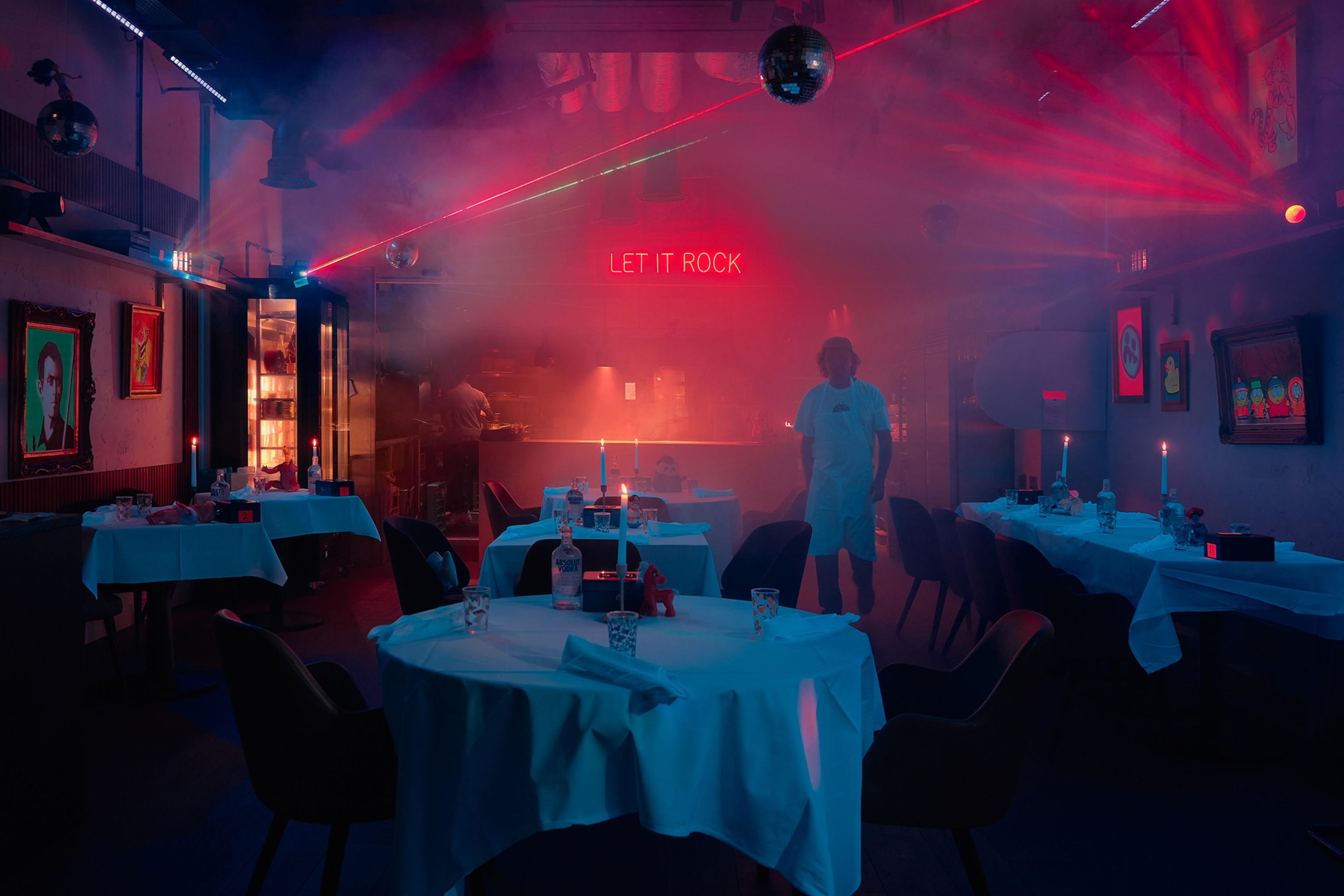 An open plan dining room of a restaurant with an open kitchen in the background and strobe lights and candles lighting up the room.