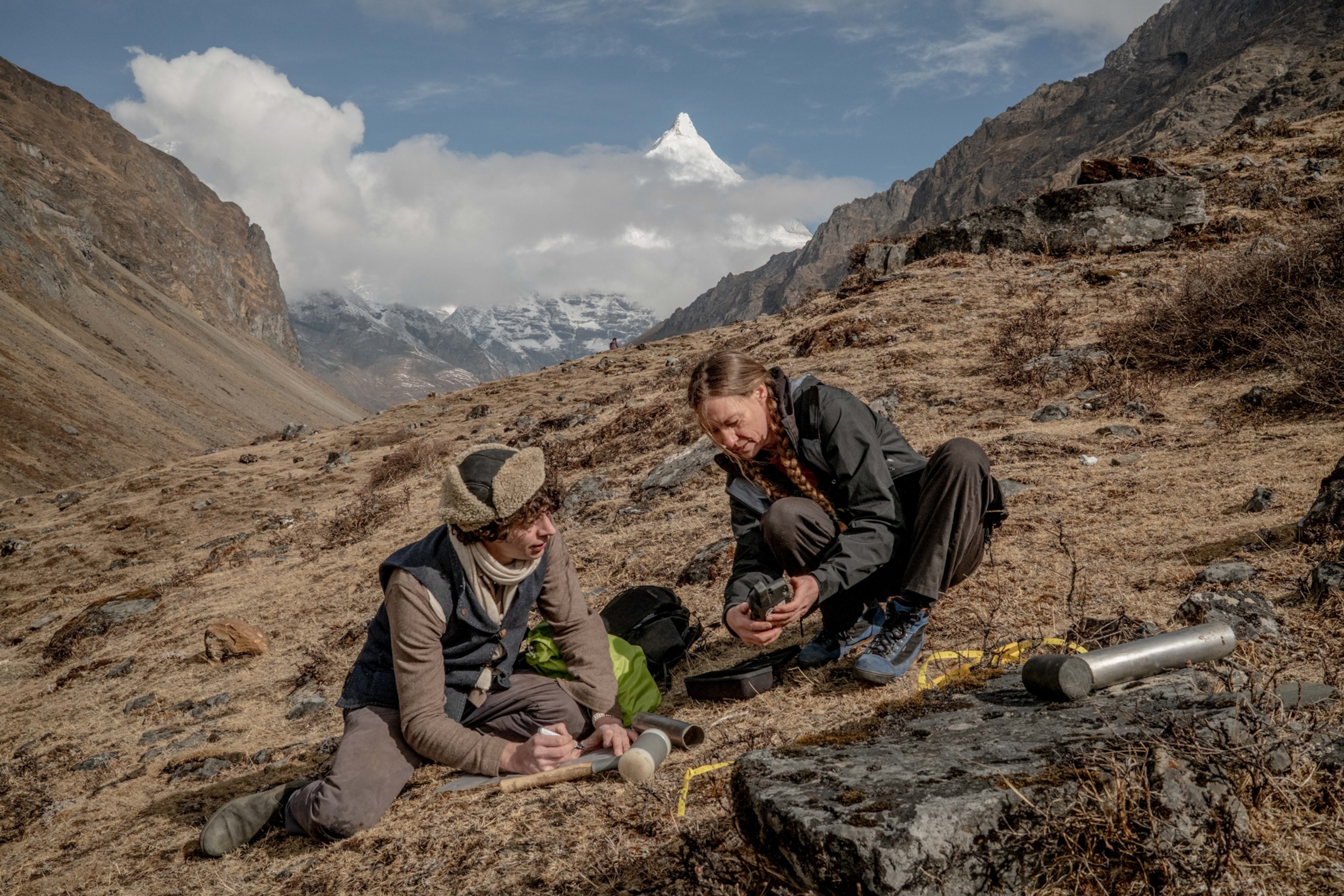 Two people kneel on a rocky mountain landscape, examining equipment. Snowy peaks are visible in the background under a partly cloudy sky.