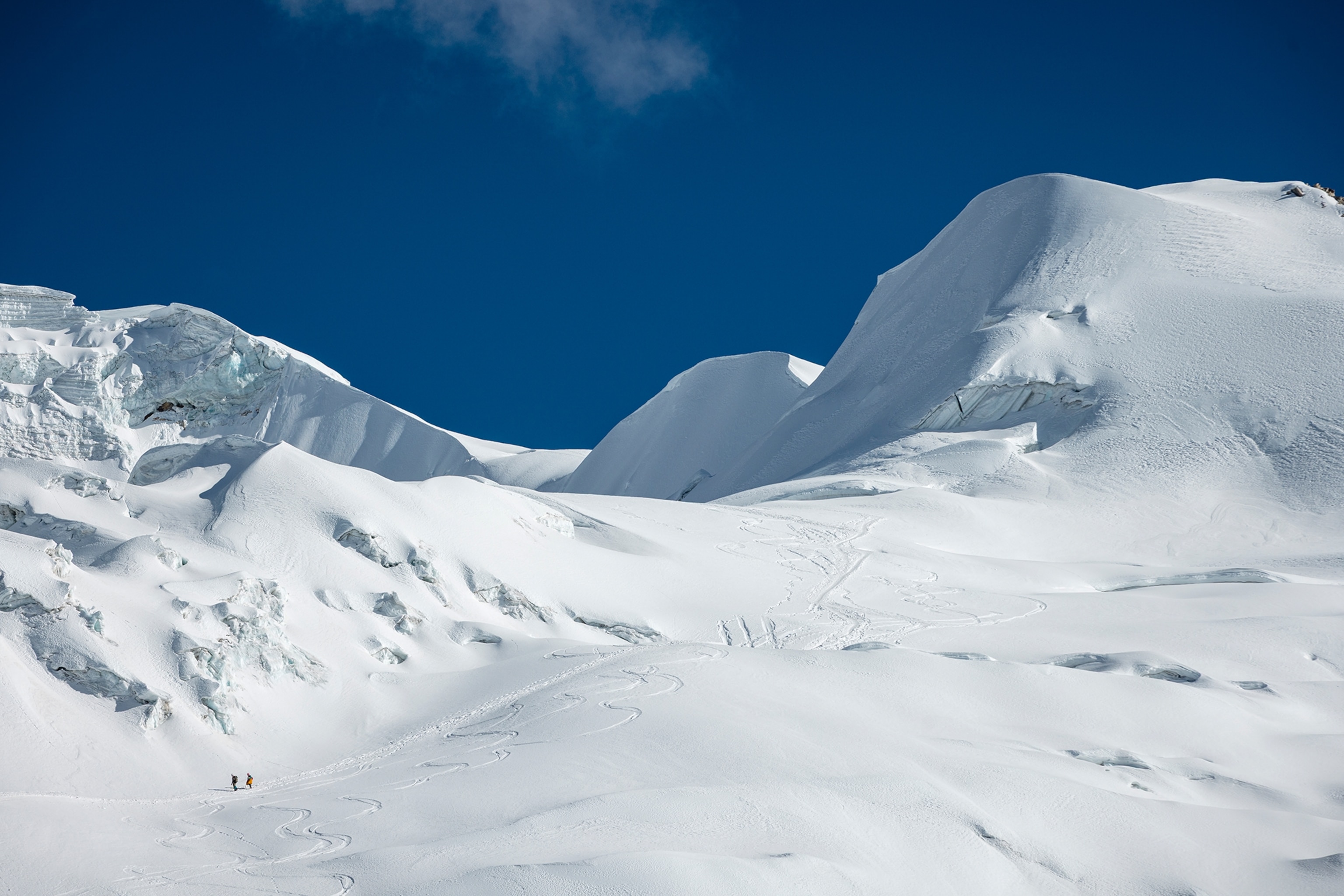 A wide, snowed-in mountain with two small climbers in the corner, making their way up to the summit.