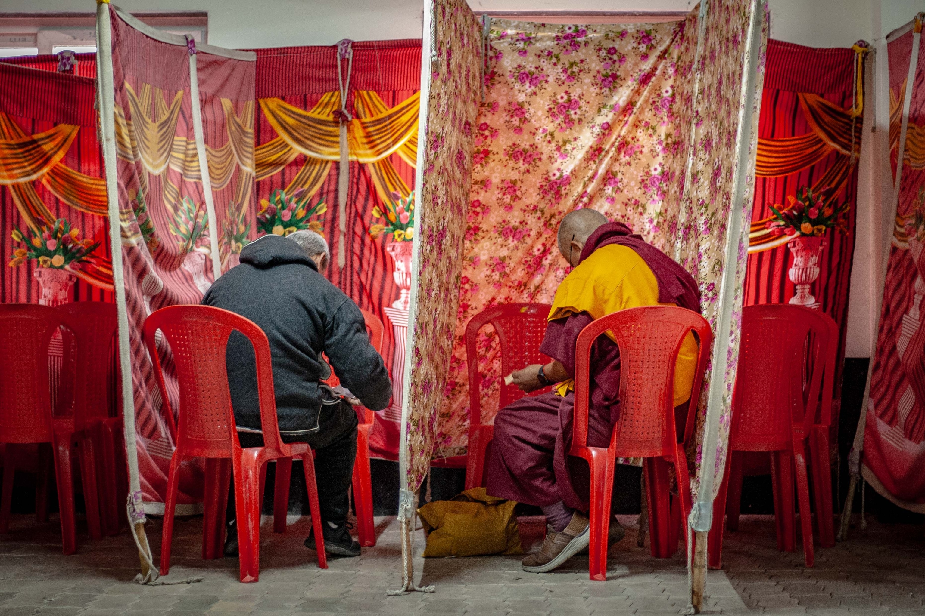 Two men sitting in red plastic chairs mark ballots at a polling station during the 2026 elections for the Tibetan government-in-exile.