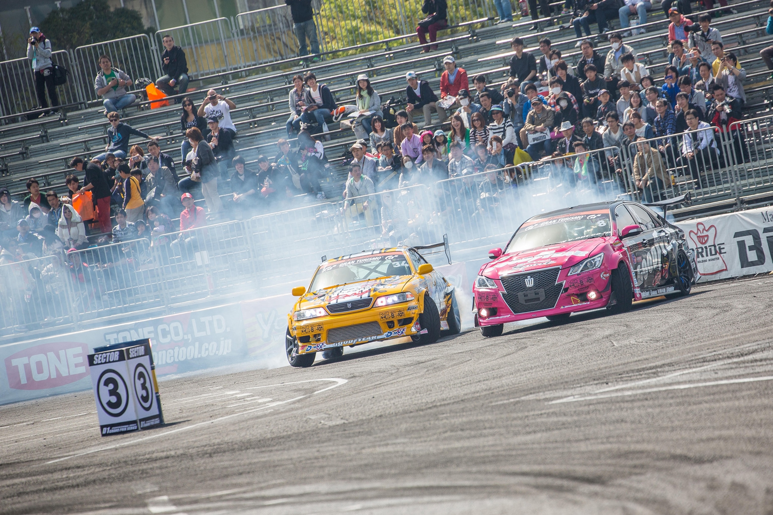 Two race cars drifting around a curve on a track with spectators in the distance.